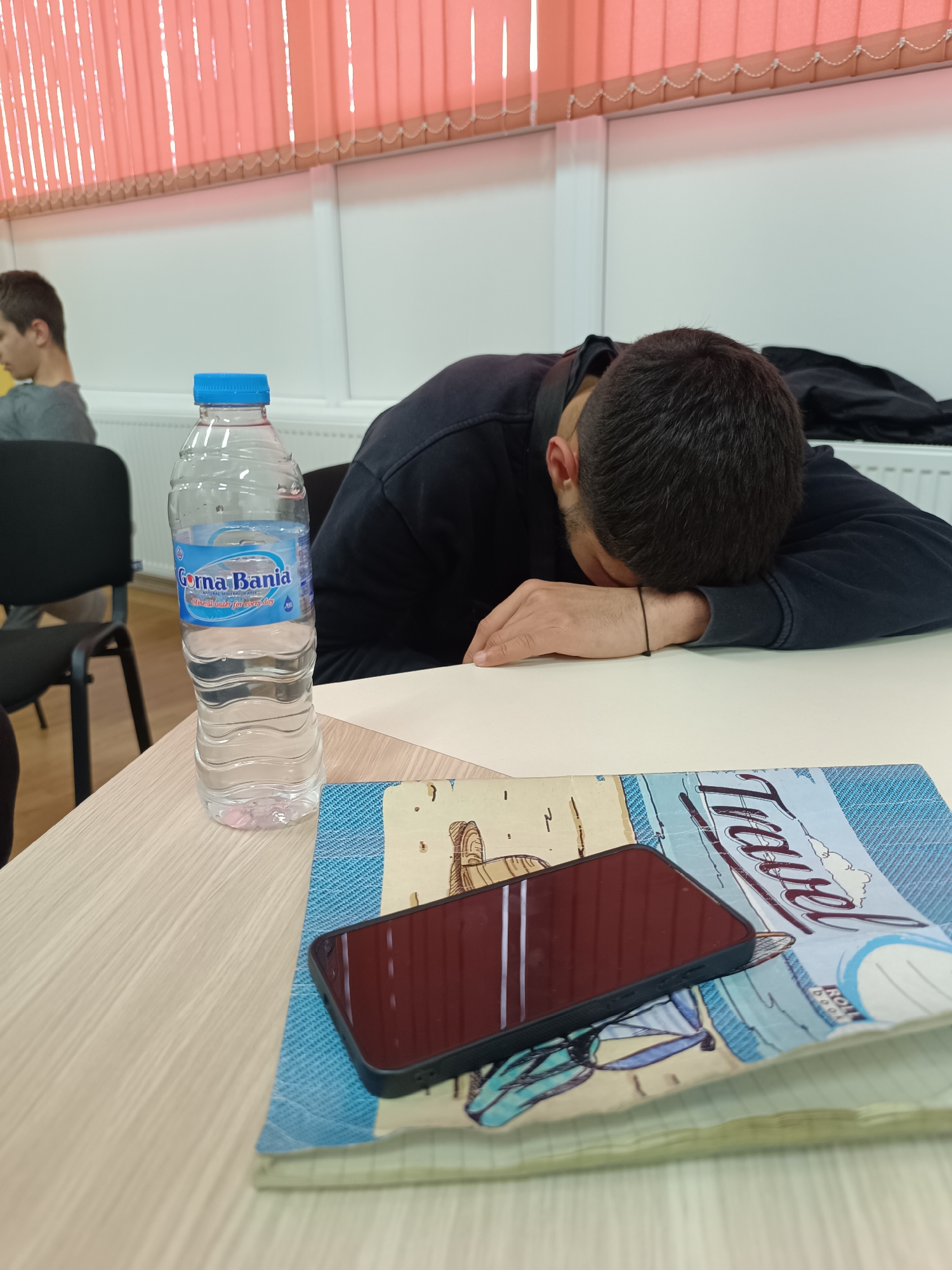 The image shows a classroom or study environment. In the foreground, a desk holds a bottle of "Gorna Bania" mineral water, a black smartphone, and a notebook with the word "Travel" visible on its cover. A dark-haired male is slumped over the desk, appearing to be sleeping. Another person is seated in the background on the left. The background includes white walls, a radiator, and orange window blinds. Given the brand of mineral water, the location could be in Bulgaria, potentially in the city of Asenovgrad.