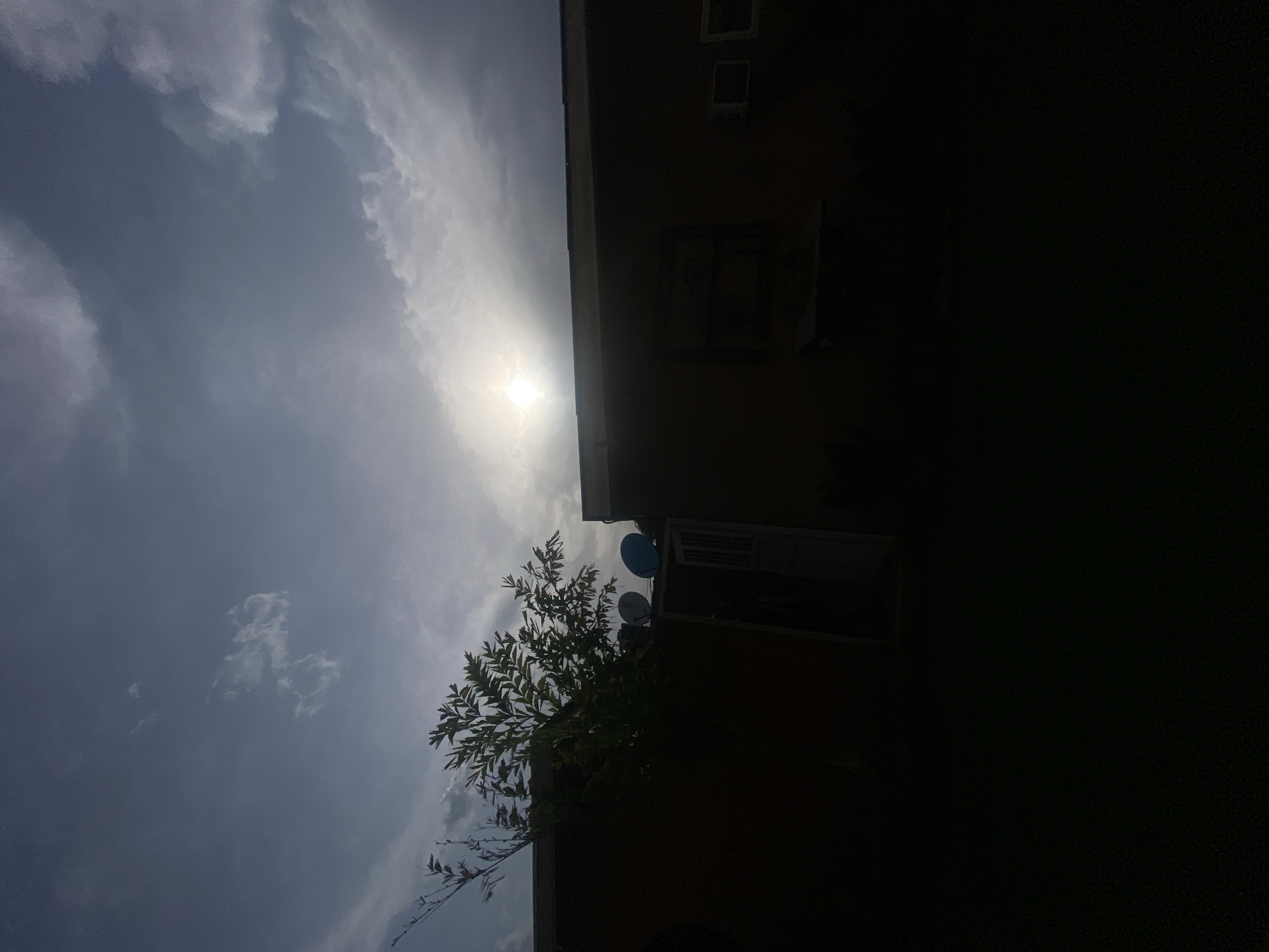 The image shows a low-angle view of a building in Barden Yana, Nigeria, under a cloudy sky with the sun shining through. The building is partially obscured by heavy shadow on the right side of the frame. Several windows are visible on the building's facade. A tree with dense foliage partially covers the building, and two satellite dishes are mounted on the exterior. The sky features varied cloud formations.