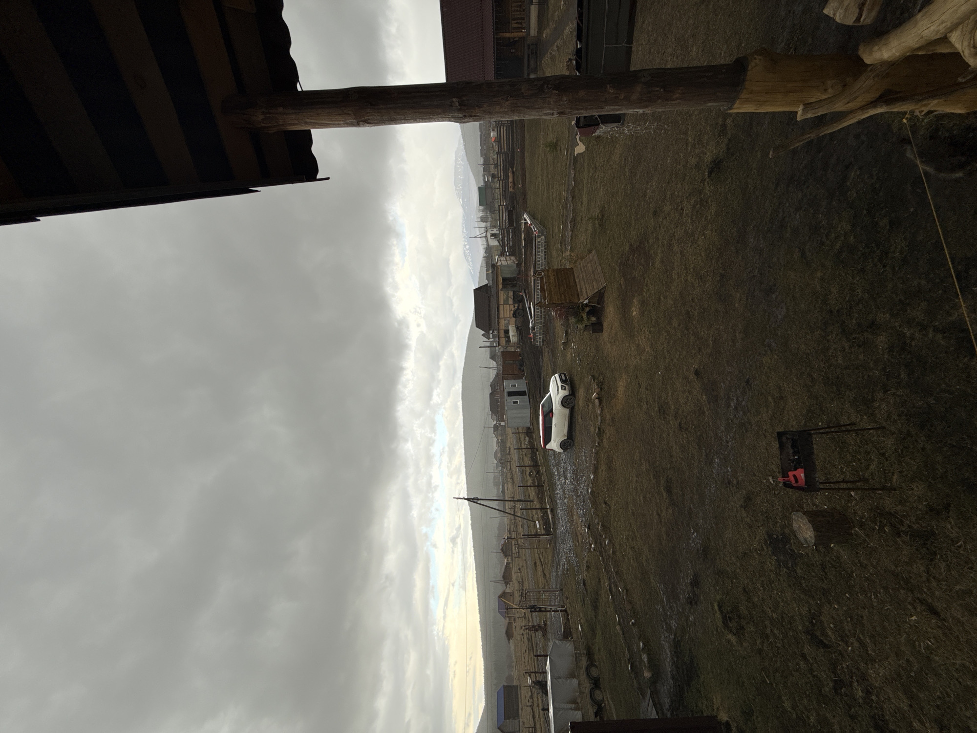 The image shows a view overlooking a landscape in what appears to be the vicinity of Kordonnyi, Russia. The sky is overcast with heavy clouds. The terrain is a mix of fields and what seems to be a small village or settlement. In the center of the frame, a white car is visible on a dirt road, parked between houses and power lines. Scattered buildings are interspersed throughout the landscape, along with outdoor furniture. In the right foreground, a grill stands on the grass. Wooden structures are visible in the upper portions of the foreground, including a roof and various posts.
