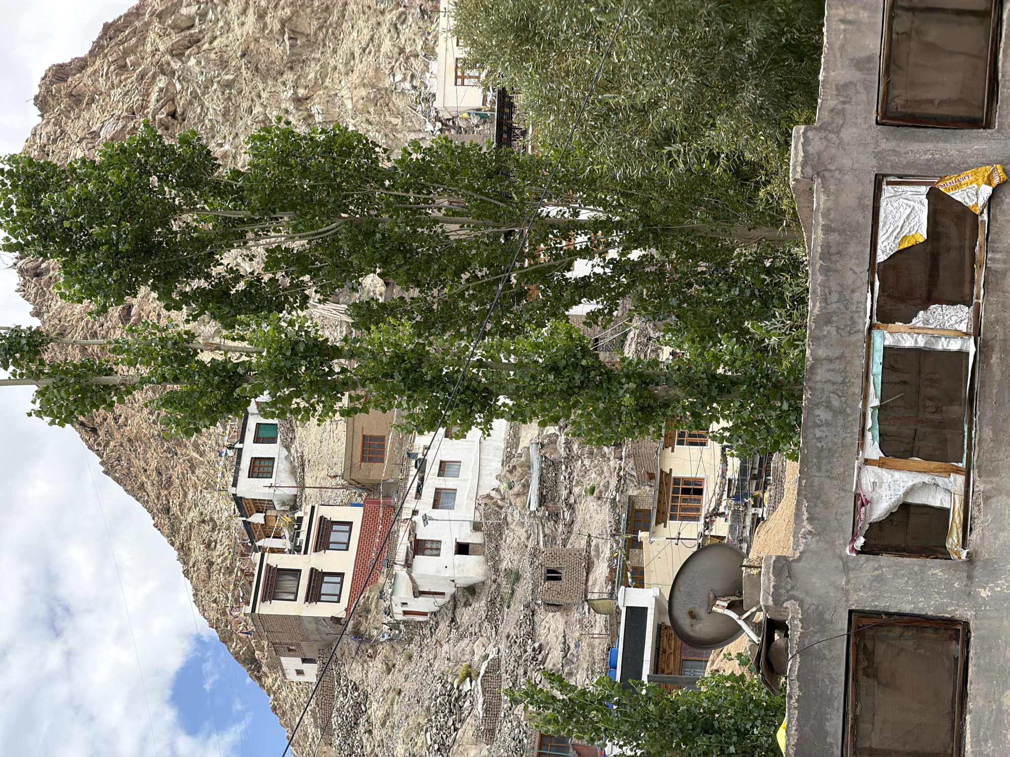 The image captures a hillside village, likely in the city of Leh, India, characterized by its arid, mountainous terrain and traditional architecture. Several multi-story buildings with white facades, wooden window frames, and flat roofs are clustered on the steep incline. Some buildings feature red brick accents and are adorned with prayer flags. The natural landscape is dominated by a rugged, rocky mountain slope that rises behind the village. Two tall, leafy trees stand prominently in the foreground, partially obscuring the view of the buildings. In the lower portion of the image, a concrete wall with several windows is visible. One of these windows is broken and partially covered with torn white and yellow material. A satellite dish is mounted on a concrete pedestal behind this wall, overlooking the village. The sky is partly cloudy, with patches of blue visible. The scene suggests a remote, possibly ancient settlement adapted to its challenging environment.
