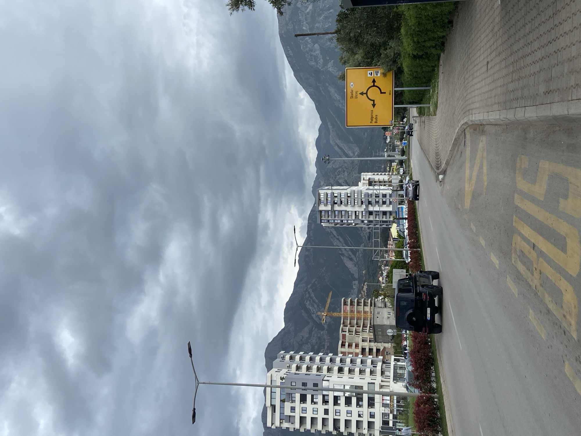 The image shows a street in Bar, Montenegro, with mountains in the background. A yellow traffic sign indicates directions to Skadar, Ulcinj, Podgorica, and Budva. Buildings of various heights line the road, including some under construction with a yellow crane visible. A black SUV is driving on the right side of the road, where yellow road markings are present. The sky is overcast with dark clouds. Sidewalks are on the right side of the frame. Streetlights are placed along the road.