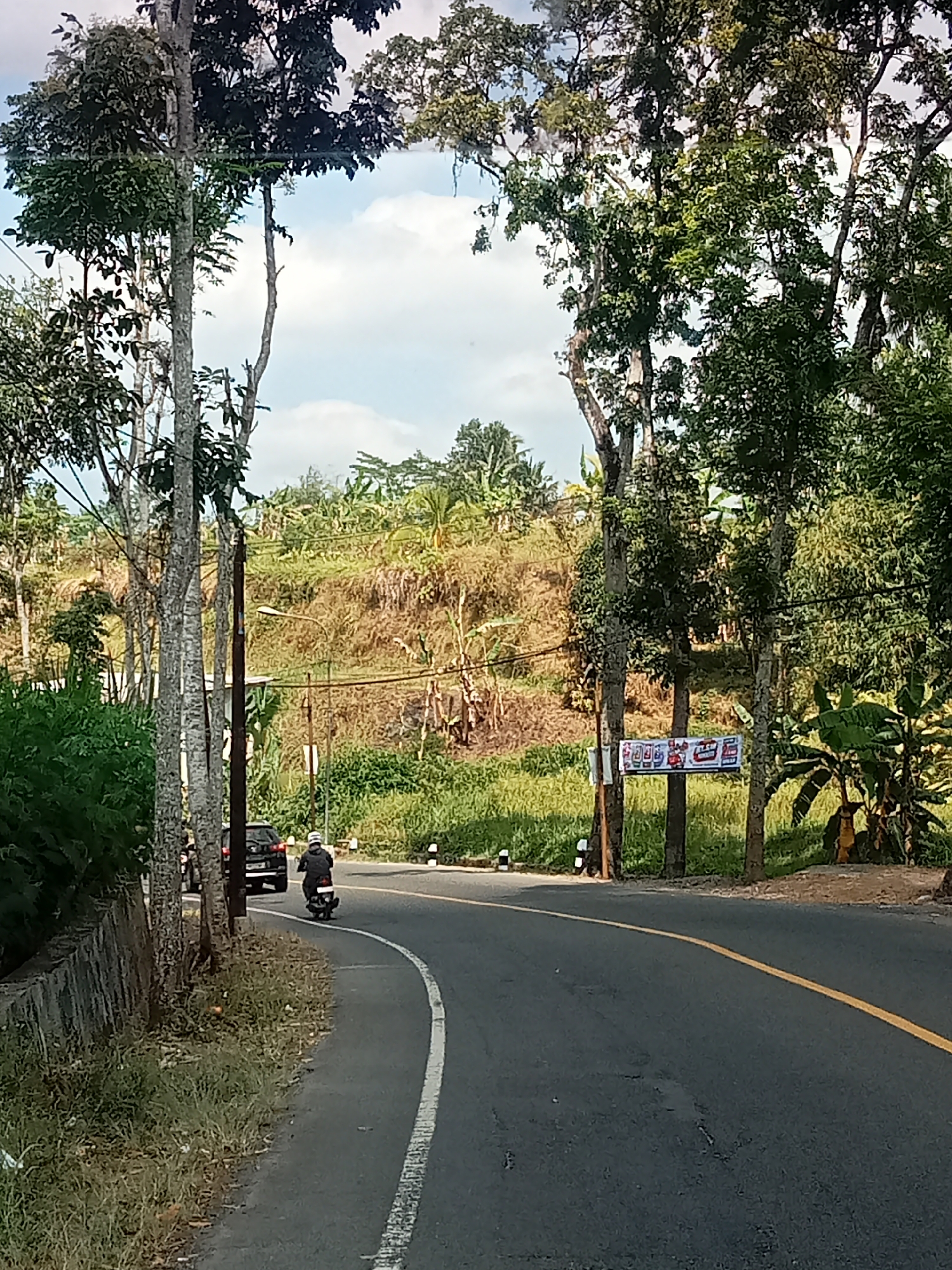 The image shows a road curving through a rural landscape in Selomerto, Indonesia. On the left side of the road, there is a grey stone wall bordering a grassy area with scattered debris. A black SUV is parked on the side of the road. A person on a motorcycle, wearing a helmet and a dark jacket, is riding away from the viewer on the right side of the road. The road has a yellow line marking the center and a white line marking the edge on the right. In the background, a grassy hillside is visible, with trees and vegetation. Two utility poles stand on the right side of the road, one with a banner attached that displays colorful text and images. The sky above is partly cloudy. The scene appears to be a typical day in a rural area of Indonesia.