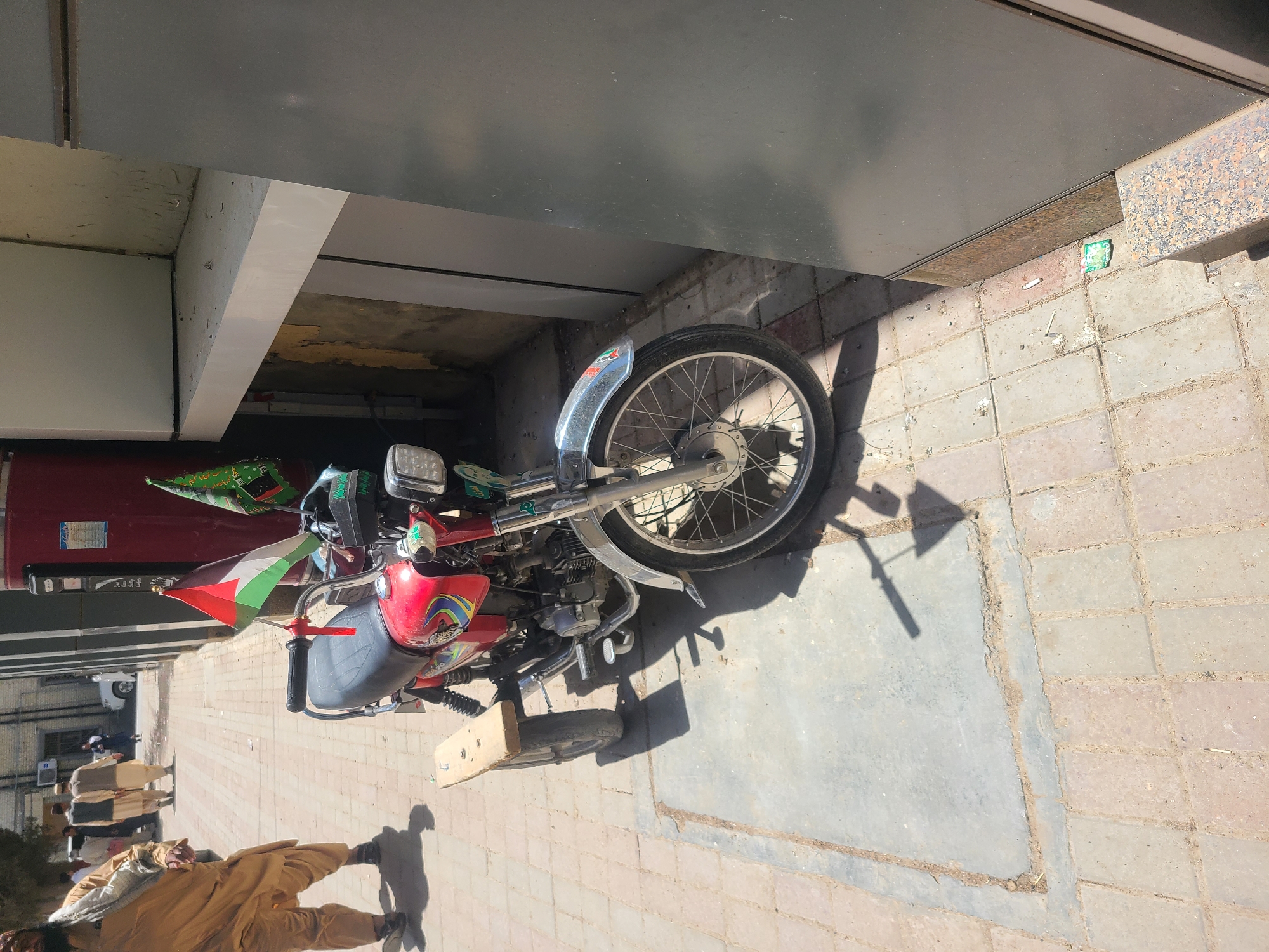 A red motorcycle adorned with a Palestinian flag is parked partially under an overhang of a building. The motorcycle's front wheel is near the edge of the overhang, casting a shadow on the paved area. A person in traditional Pakistani clothing is walking in the foreground. Other individuals are visible in the background on the left. The setting appears to be a city street in Quetta City Tehsil, Pakistan.