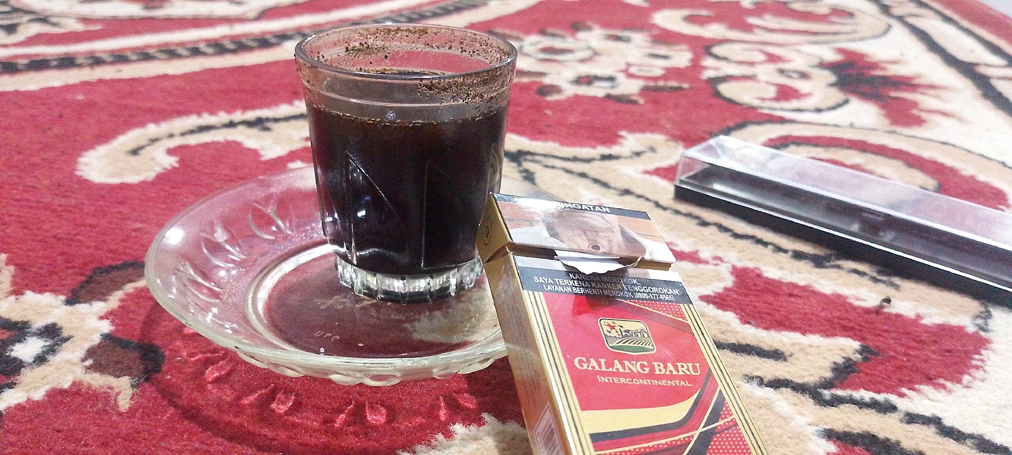 A close-up, low-angle, outdoor, daytime shot shows a glass of dark liquid, possibly coffee, on a clear glass saucer, situated on a red and tan patterned rug. The saucer has an etched decorative pattern around its rim. To the right of the glass is an open pack of "GALANG BARU INTERCONTINENTAL" cigarettes, which features a graphic image of a man's face and text in Indonesian warning about health risks. A clear plastic cigarette case is visible behind the cigarette pack. The scene is set in what appears to be an outdoor area, suggested by the rug and the presence of the cigarette pack, typical of informal social gatherings in Indonesian locations such as Oro-oro Ombo. The lighting is natural, creating subtle shadows on the rug.