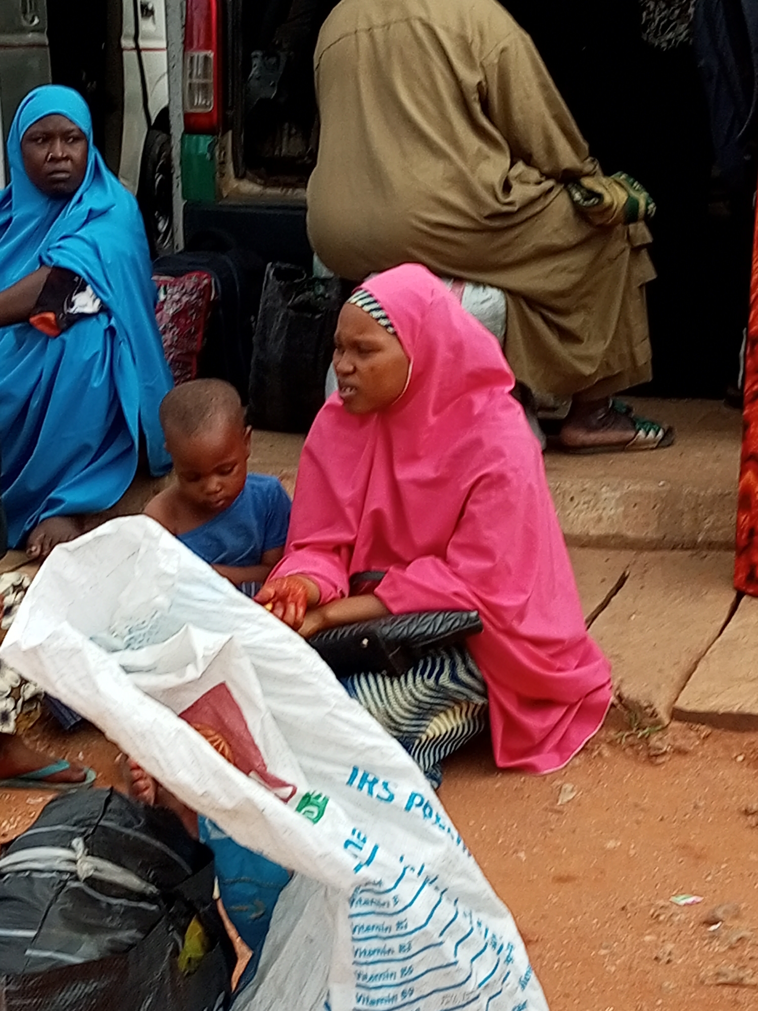 In Gombe, Nigeria, at a dusty outdoor location, possibly a market or transportation hub, several individuals are gathered around a white, open sack. A woman in a bright pink hijab sits on the ground, her hands stained with what appears to be henna or dye, and she is interacting with a young child. The child, wearing a blue shirt, looks down with a somber expression. Another woman, dressed in a blue hijab, is seated to the left, her gaze directed forward. In the background, a person in a tan garment is partially visible, appearing to be seated on steps. The environment is characterized by reddish-brown earth and scattered debris. The white sack in the foreground displays text, including "IRS PO" and lists of "Vitamin B" concentrations, suggesting it might contain nutritional supplements or animal feed. Other bags and what seem to be personal belongings are scattered around the area, contributing to a sense of a temporary gathering or transit point.
