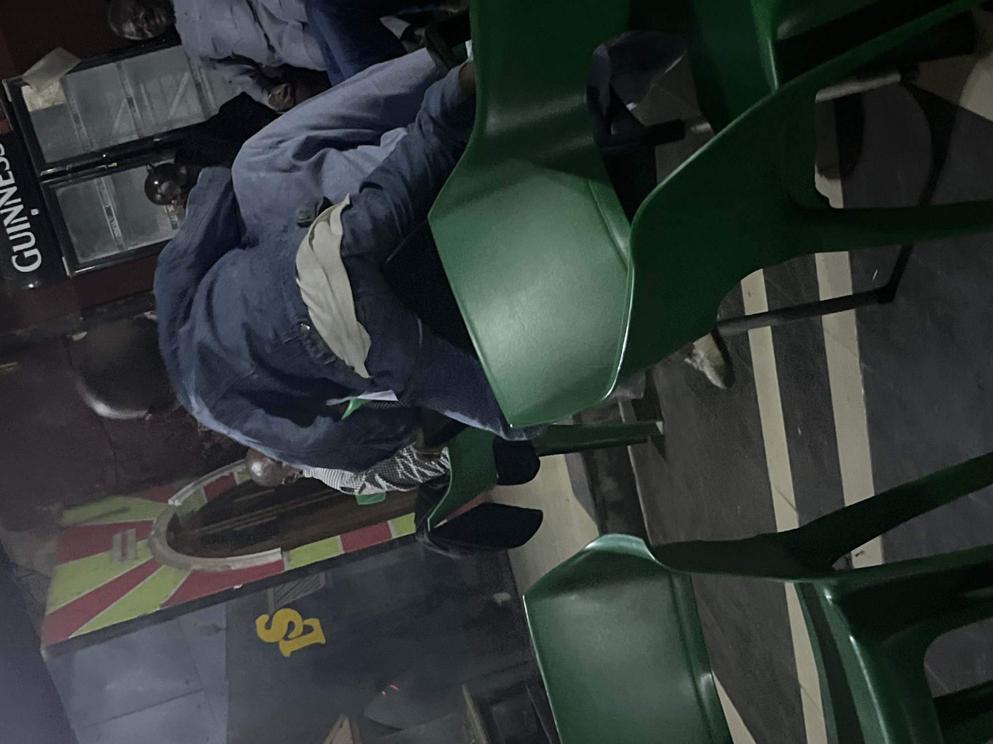 A low-angle, medium shot captures an indoor scene in Jos, Nigeria, where individuals are seated in green plastic chairs. In the foreground, a child wearing a denim jacket and jeans is perched on the back of a chair, facing away from the camera. The child's right leg is extended, resting on another person's lap. Behind the child, another person in a blue shirt is seated. Further in the background, a Guinness refrigerator is visible, with three men standing or sitting near it. To the left of the refrigerator, there is a signage that appears to read "LS" in yellow letters against a dark background, with red and green stripes framing an archway. The lighting is dim, suggesting an evening setting. The floor is tiled with a pattern of black and light-colored stripes.