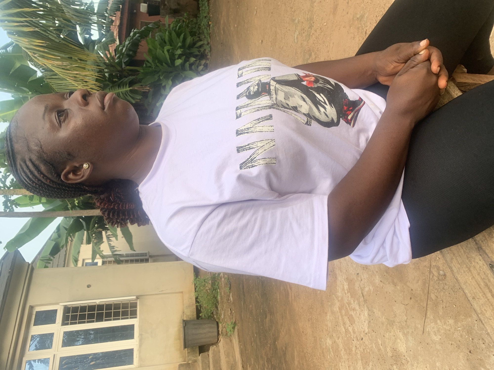 The image depicts a young Black woman sitting outdoors in what appears to be a residential area. She has her hair in braids and is wearing a white t-shirt with a graphic print and black leggings. The t-shirt has the word "NINJA" in large block letters, with a stylized depiction of a person in a hat and jacket below it. Her hands are clasped in her lap.
The background includes a building with large windows and a tan-colored facade, suggesting a home or similar structure. There are also lush green plants, including what appear to be banana leaves, and a dirt-covered ground with some scattered debris. The setting suggests a warm climate, consistent with Akwanga, Nigeria. The overall impression is of a casual, everyday scene.