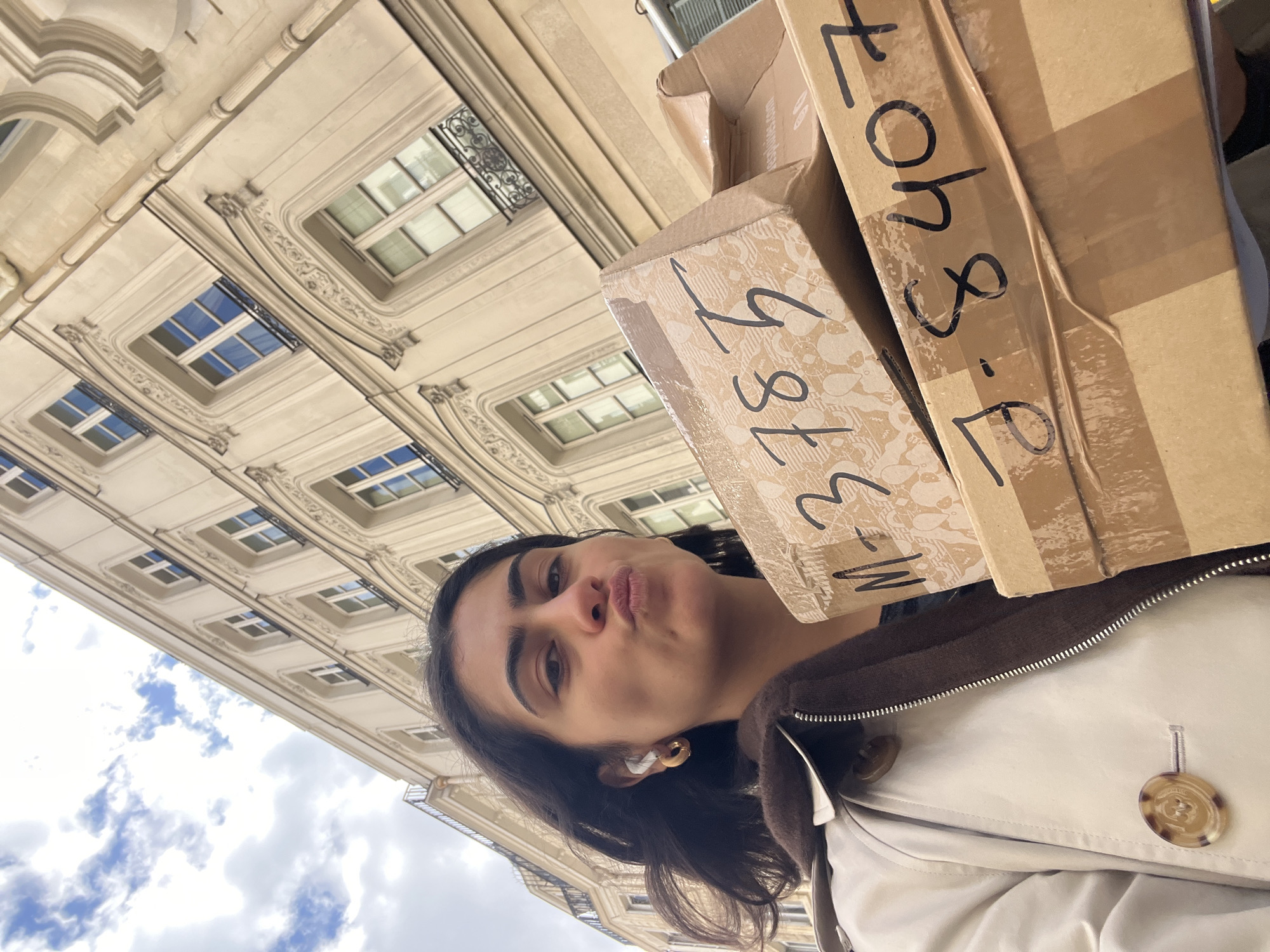 A low-angle, outdoor selfie captures a woman holding several cardboard boxes against a backdrop of Parisian architecture and a cloudy sky. The woman has dark hair, fair skin, and is wearing wireless earbuds. She is making a kissing face at the camera, with her eyes directed toward the lens. Her attire consists of a white jacket with brown accents and large, decorative buttons.
The cardboard boxes are stacked atop one another, with shipping labels and handwritten markings visible on their exteriors. The markings include numbers and possibly shipping codes.
The background features a grand Parisian building with ornate window frames, classical architectural details, and a pale facade. Above the building, a partially cloudy sky is visible, suggesting a bright day with scattered clouds. The perspective is tilted, giving the impression that the photo was taken while looking up at the buildings.