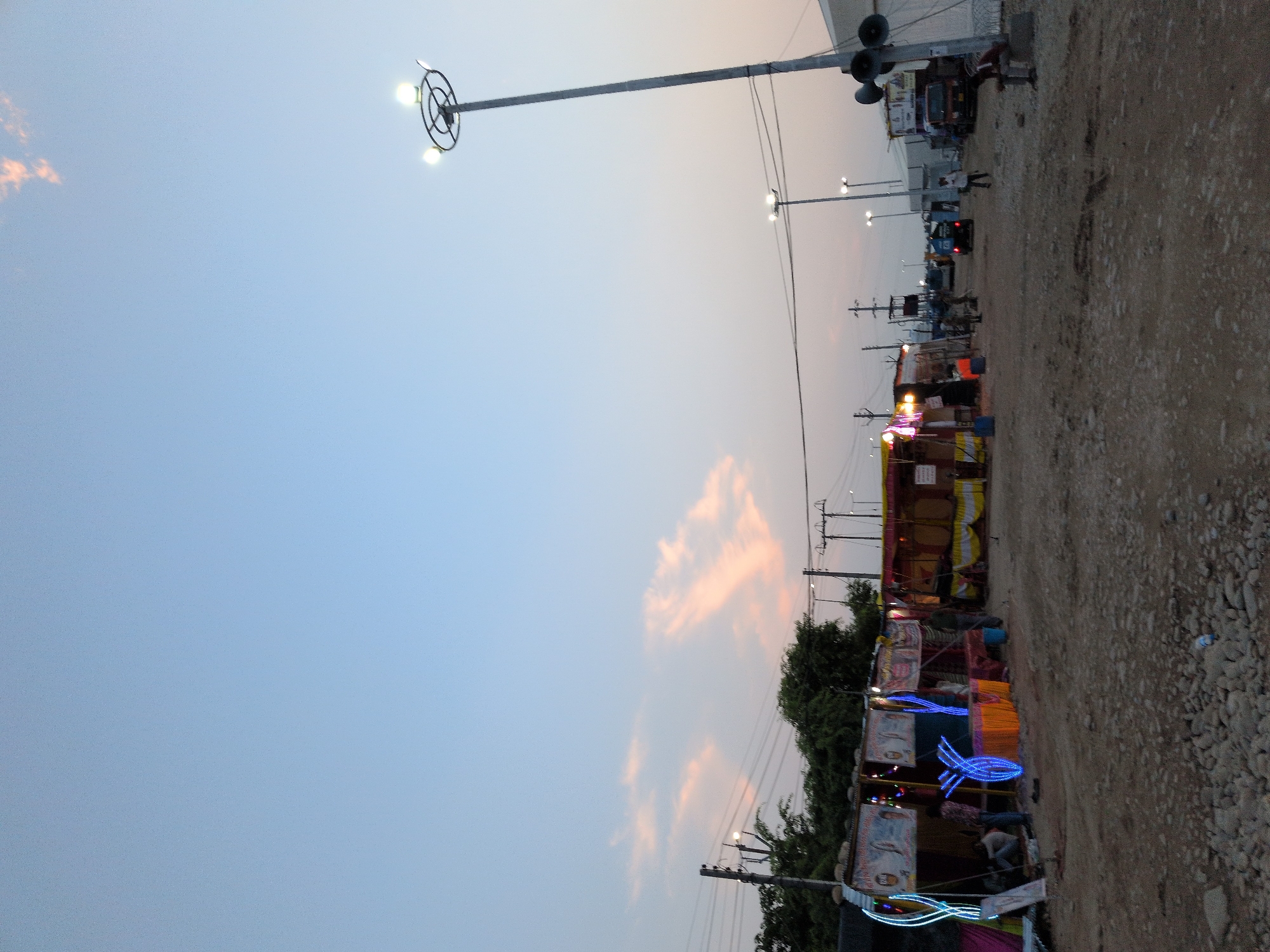This is a full-frame, eye-level, outdoor, ground-level shot taken in Jammu, India. The scene depicts a dusty, unpaved road lined with temporary stalls or shops, some adorned with festive lighting. In the foreground, the ground is covered with small pebbles and dirt, with a plastic bottle visible. Several stalls are set up along the left and center of the image, featuring fabric awnings and decorated with colorful lights, including blue neon-like strips. Some people are seen near the stalls, with one individual sitting and another standing.
Further down the road, more stalls and structures are visible, along with vehicles, including what appears to be a truck and a smaller utility vehicle. Utility poles with electrical wires crisscross the scene, and a tall light pole with multiple lamps stands prominently on the right side of the image. A large speaker system is mounted on the light pole. The sky above is a pale blue with wispy pinkish clouds, indicating it is either dawn or dusk. The overall impression is of a temporary marketplace or gathering place, possibly during a festival or event.