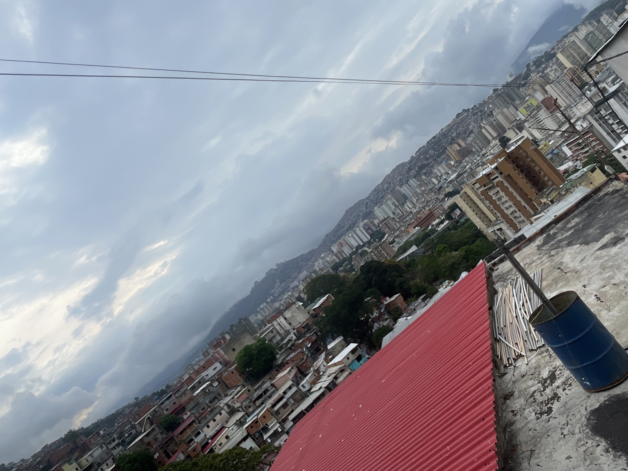 The image captures a wide, diagonal view of a dense urban landscape, likely Caracas, Venezuela, under an overcast sky. The perspective is from a high vantage point, possibly a rooftop, looking across the city towards distant mountains that are heavily populated with buildings.
In the immediate foreground, a vibrant red, corrugated metal roof stretches across the lower-left portion of the frame. Adjacent to it, on a rough concrete surface in the lower right, stands a blue metal barrel, partially filled with several white and grey pipes or rods. Two dark, thin power lines traverse the upper third of the frame against the sky.
The sky is heavily clouded with varying shades of grey and white, indicating an overcast day. The cityscape extends far into the distance, showcasing a mix of multi-story buildings, including modern high-rises and older, more densely packed residential structures. These buildings climb the slopes of the surrounding mountains, which form the backdrop of the scene. Areas with more modest, closely built homes, characteristic of informal settlements or traditional barrios, are also visible.
A yellow and blue sign displaying the word "Daka" can be seen on a building in the middle distance. No people are visible, and the scene depicts a static overview of the urban sprawl against its natural, mountainous setting.