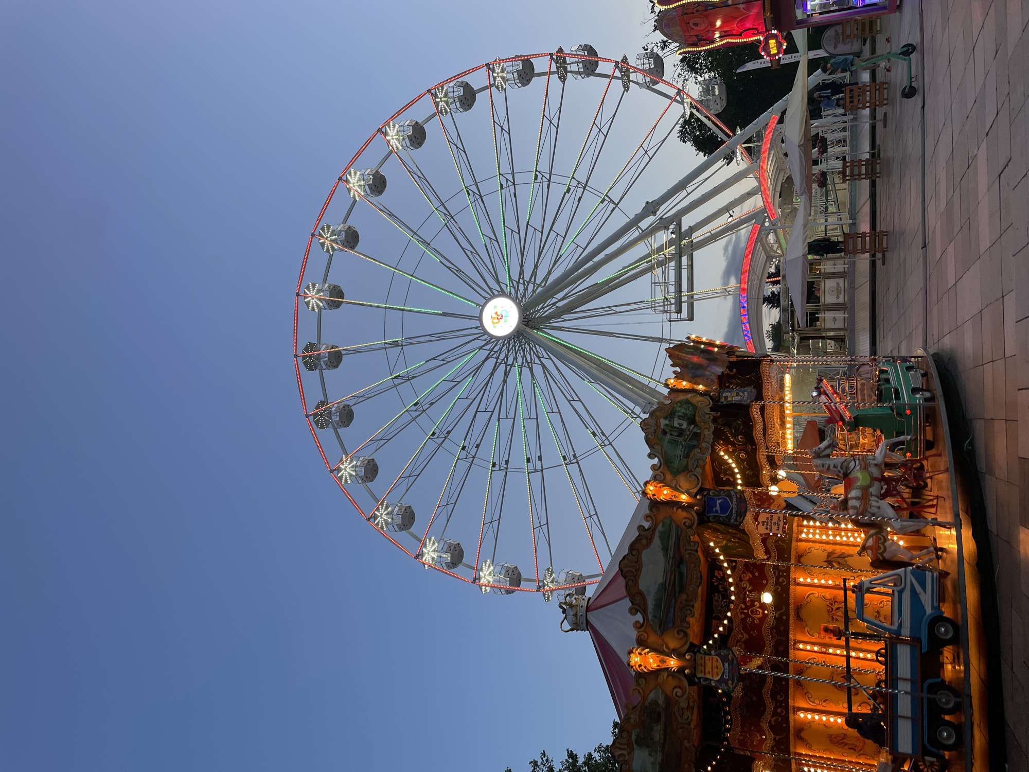 The image displays a lively amusement park scene captured at dusk or early evening in Sofia, Bulgaria. The sky is a clear, deep blue, providing a contrasting backdrop for the illuminated attractions.
Dominating the left side of the frame is a large Ferris wheel, its white or light-colored frame brightly lit. Red lights line its outer rim, while green and white lights extend along its spokes towards a central hub that features a glowing white circle with a colorful, unidentifiable logo. Several gondolas are visible, some appearing to carry passengers enjoying the view.
In the foreground, towards the right, a traditional carousel is brightly illuminated with warm yellow and orange lights. It features classic carousel figures, including white and brown horses, as well as a distinct blue and white truck-like ride, upon which a person, likely a child, can be seen. The carousel is adorned with ornate decorative panels, though any text on them is not clearly legible.
In the background and midground, other fair stalls or structures are visible, some emitting red light. Paved ground, resembling cobblestones or tiles, covers the bottom and right portions of the scene. Seating areas with tables, chairs, and a white umbrella suggest spaces for visitors to relax. Dark green foliage from trees is visible in the background, adding a touch of nature. The overall atmosphere is festive and joyful, characteristic of an evening fair.