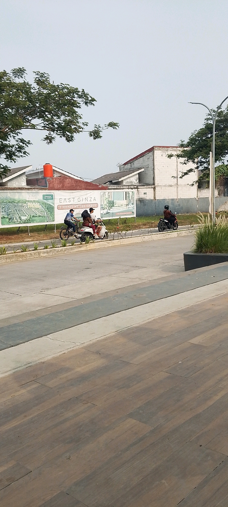 In Satriamekar, Indonesia, a scene unfolds on a paved road adjacent to a construction site. Three individuals are on motorcycles. Two people ride on one scooter, and a single person is on another motorcycle further down the road. Behind them, large billboards display real estate advertisements. The sky is overcast, and the surrounding area features trees and newly constructed buildings. The foreground is dominated by a wooden walkway, suggesting a public space or park area.