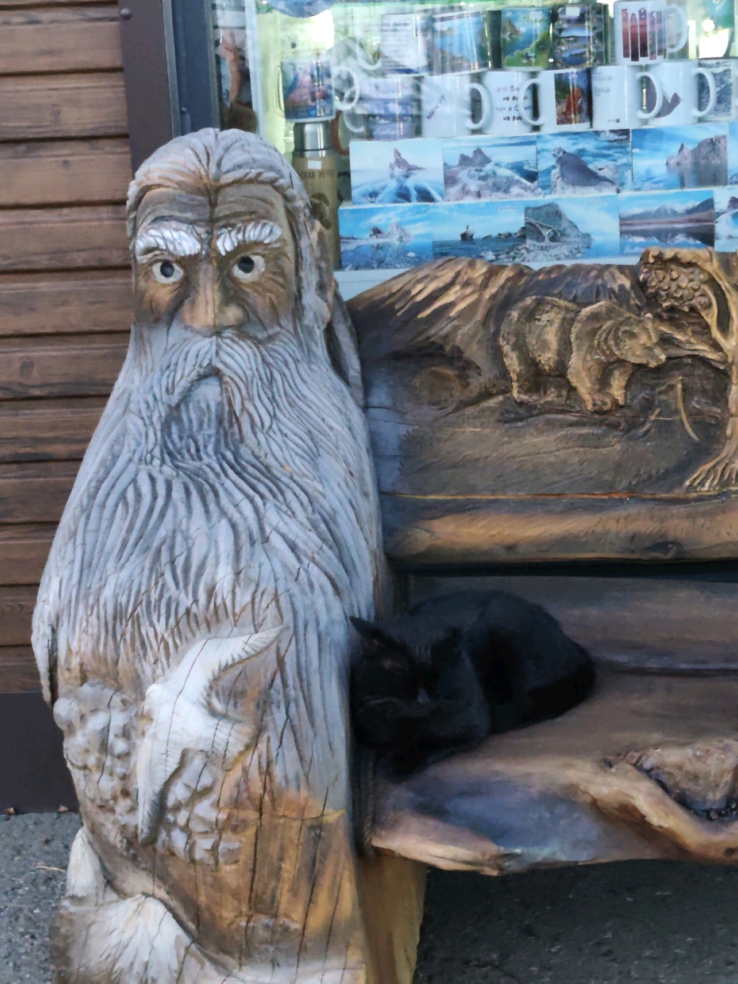 A carved wooden sculpture of an old man with a long beard and a bird in relief on its chest sits next to a wooden bench carved with a bear and a tree. A black cat is curled up asleep on the bench. In the background, through a glass window, shelves are stocked with ceramic mugs and framed pictures. The sculpture and bench are positioned in front of a wooden building. The scene appears to be a tourist-oriented setting, likely in Listvyanka, Russia, given the context of the prompt.