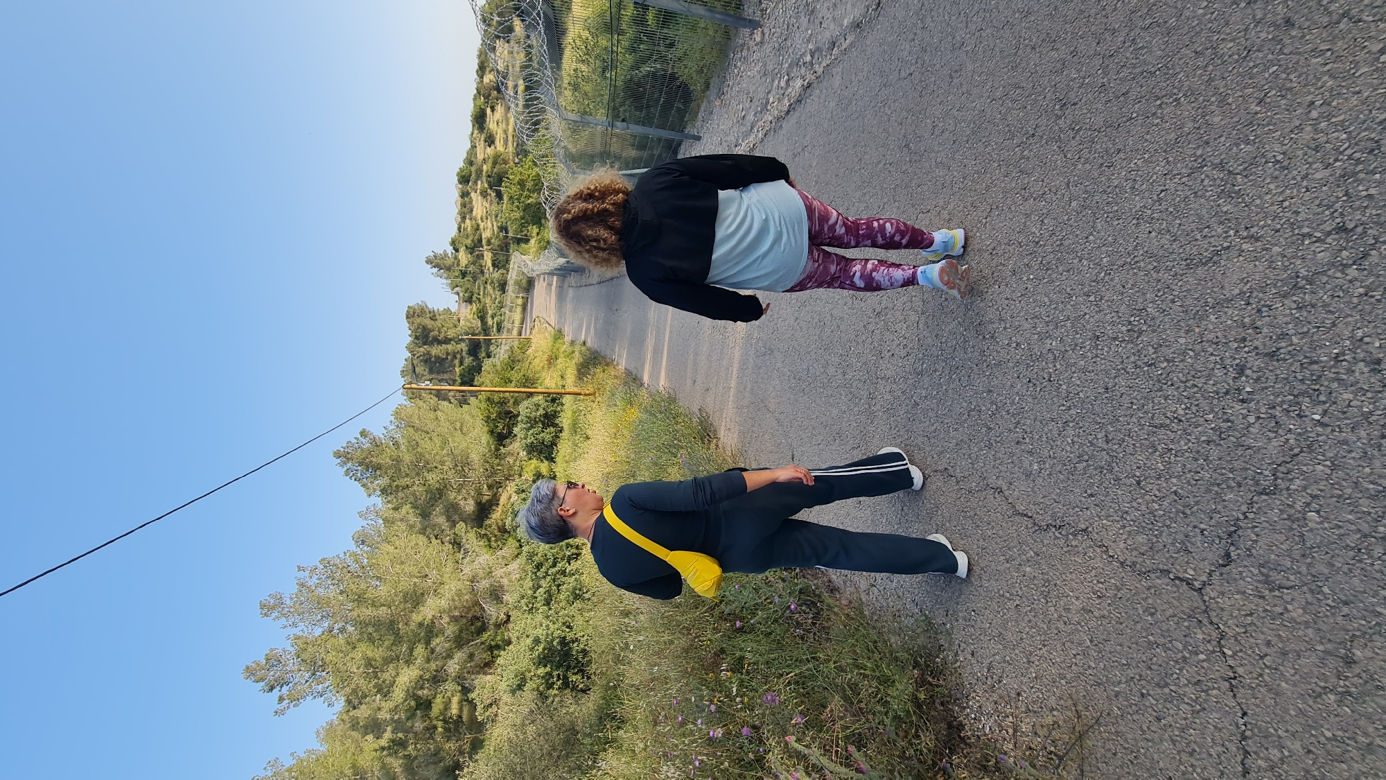 Two people are walking on a paved road in Lachish Regional Council, Israel. One person is wearing black pants with a white stripe, a black long-sleeve shirt, and a yellow bag. The other person is wearing a white t-shirt, a black jacket, and patterned leggings. There is greenery and barbed wire on the side of the road. The sky is clear and blue.