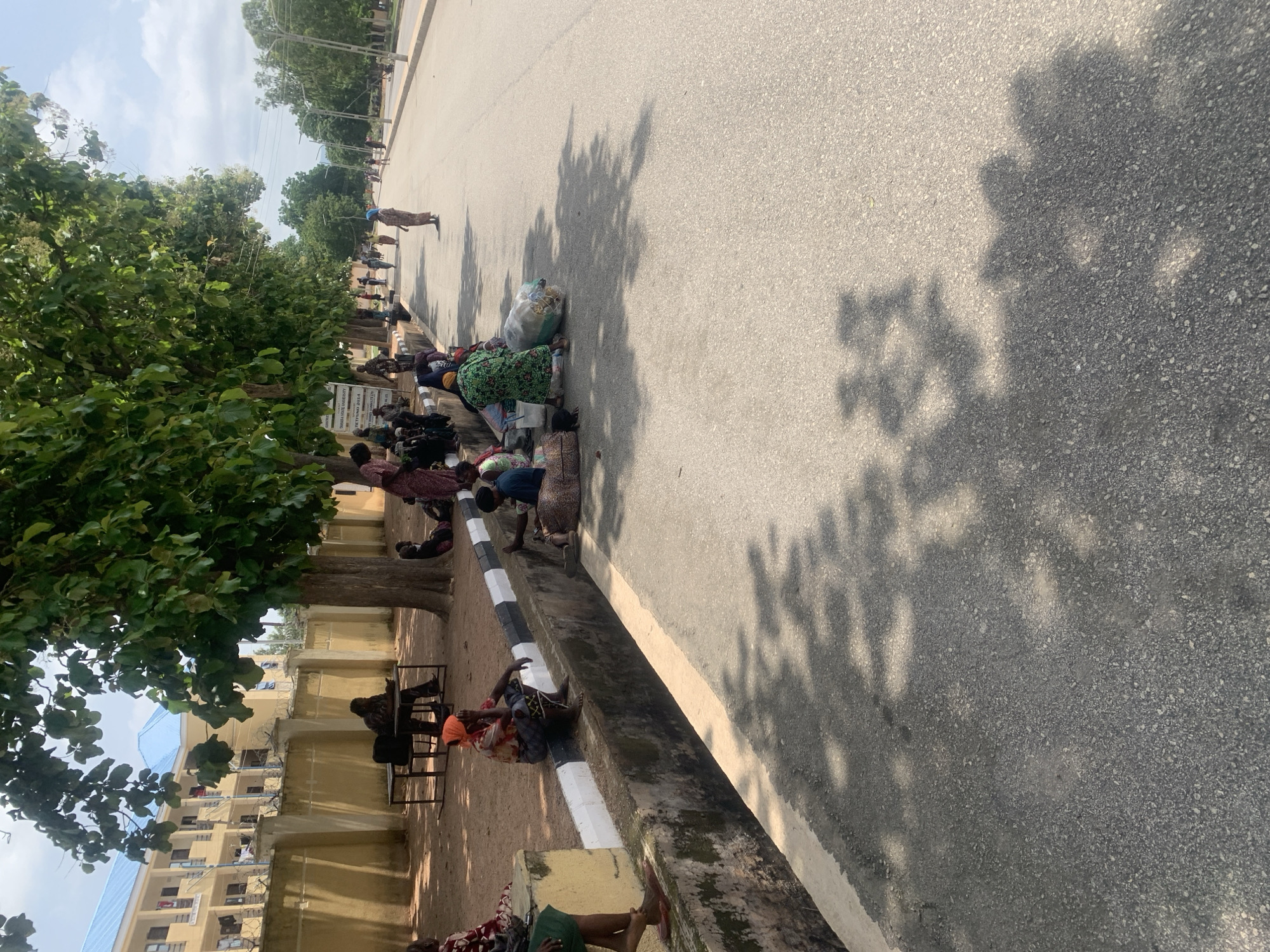 This image depicts an outdoor scene on a street in Jalingo, Nigeria, likely on the campus of Taraba State University, as suggested by a partially visible sign in one of the cropped images. The foreground shows a paved road with shadows of trees cast across it, indicating it is daytime. Along the left side of the road is a concrete drainage channel with a black and white striped border. A group of people, predominantly women dressed in traditional Nigerian attire, are seated or gathered along the edge of this channel and on the roadside.
Some individuals are sitting on the ground, while others are perched on the concrete border. Several large bags or bundles are also present, suggesting they may be waiting for transport or are engaged in some form of trade or activity. In the background, across the street, there are buildings with light-colored walls and blue roofs, and a tall fence. Trees line the side of the road where the people are gathered, providing shade. The overall atmosphere appears to be one of a public space where people are congregating, possibly waiting or resting.