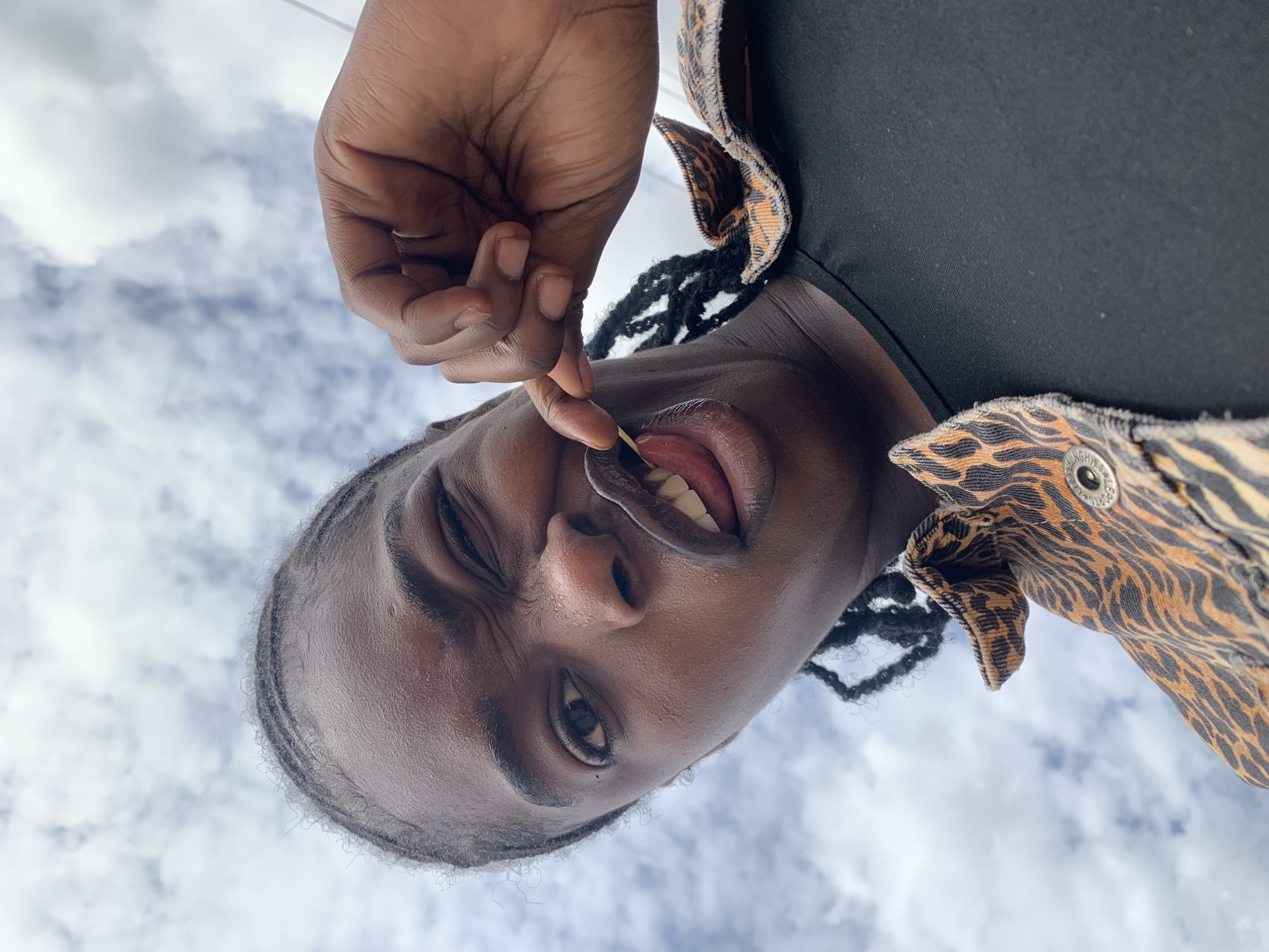 The scene is set in New Stadium, Nigeria. A single individual is captured from a low-angle perspective, positioned against an overcast sky with scattered white and grey clouds. The person has a dark complexion and braided hair, visible along the forehead and sides. They are looking towards the camera, winking with their right eye while their left eye remains open. Their mouth is open, with the tongue partially extended. A slender, light-colored object, resembling a toothpick or small stick, is held between their upper lip, tongue, and the thumb and index finger of their right hand. The individual wears a black crew-neck shirt beneath a collared jacket featuring an orange and black animal-print pattern (resembling tiger or zebra stripes), visible across the shoulders and upper chest.