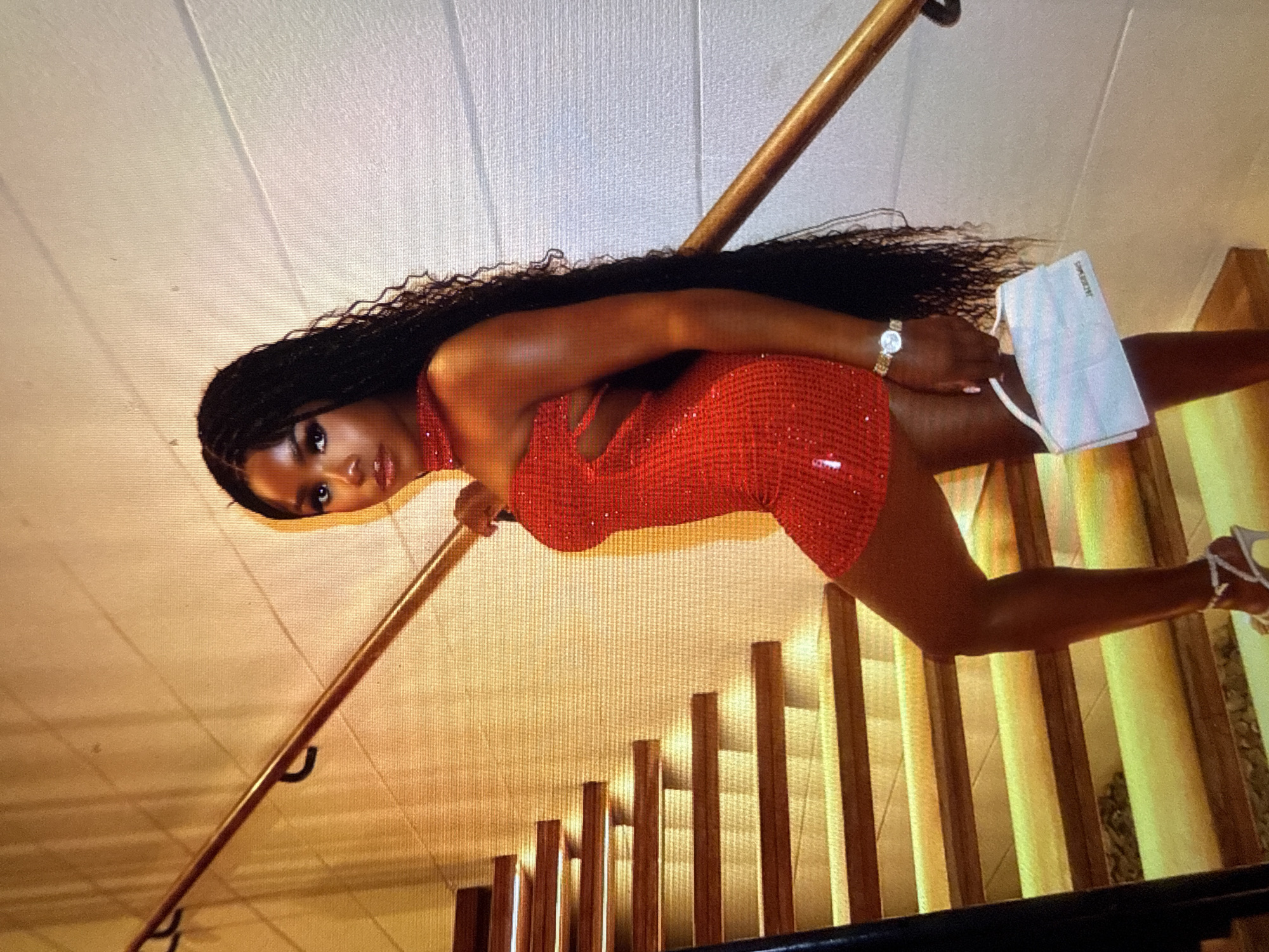 A young Black woman with long, dark braided hair stands on a staircase, looking at the camera. She is wearing a sparkling red dress with cutouts on the chest and a high neckline. She has on a watch on her left wrist and is holding a light blue face mask in her right hand. She is wearing strappy, white high-heeled shoes. The staircase has wooden steps and a wooden banister. The walls are painted a light color, and there are shadows cast by the lighting. The scene appears to be indoors, possibly in a home. The overall mood is confident and celebratory, suggesting a special occasion. The visible text on the face mask reads "Jacquemus."