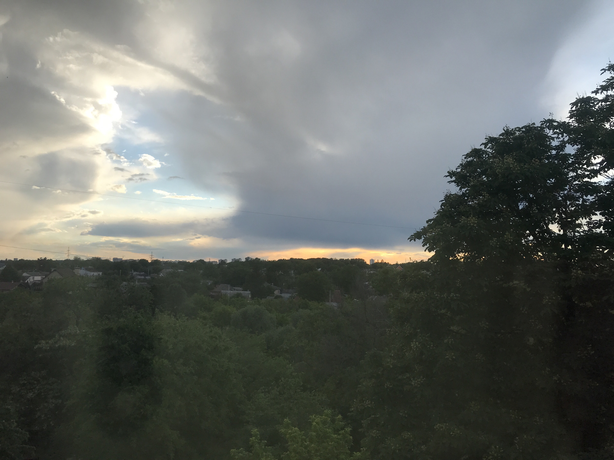 The image shows a view overlooking a cityscape partially obscured by dense trees. A line of buildings is visible in the middle distance, with power lines cutting across the sky. Above the buildings is a dynamic sky filled with gray clouds, with a break in the clouds showing a patch of blue sky and hints of sunset. The right side of the frame is dominated by a tree. The location is possibly Bezliudivka, Ukraine.