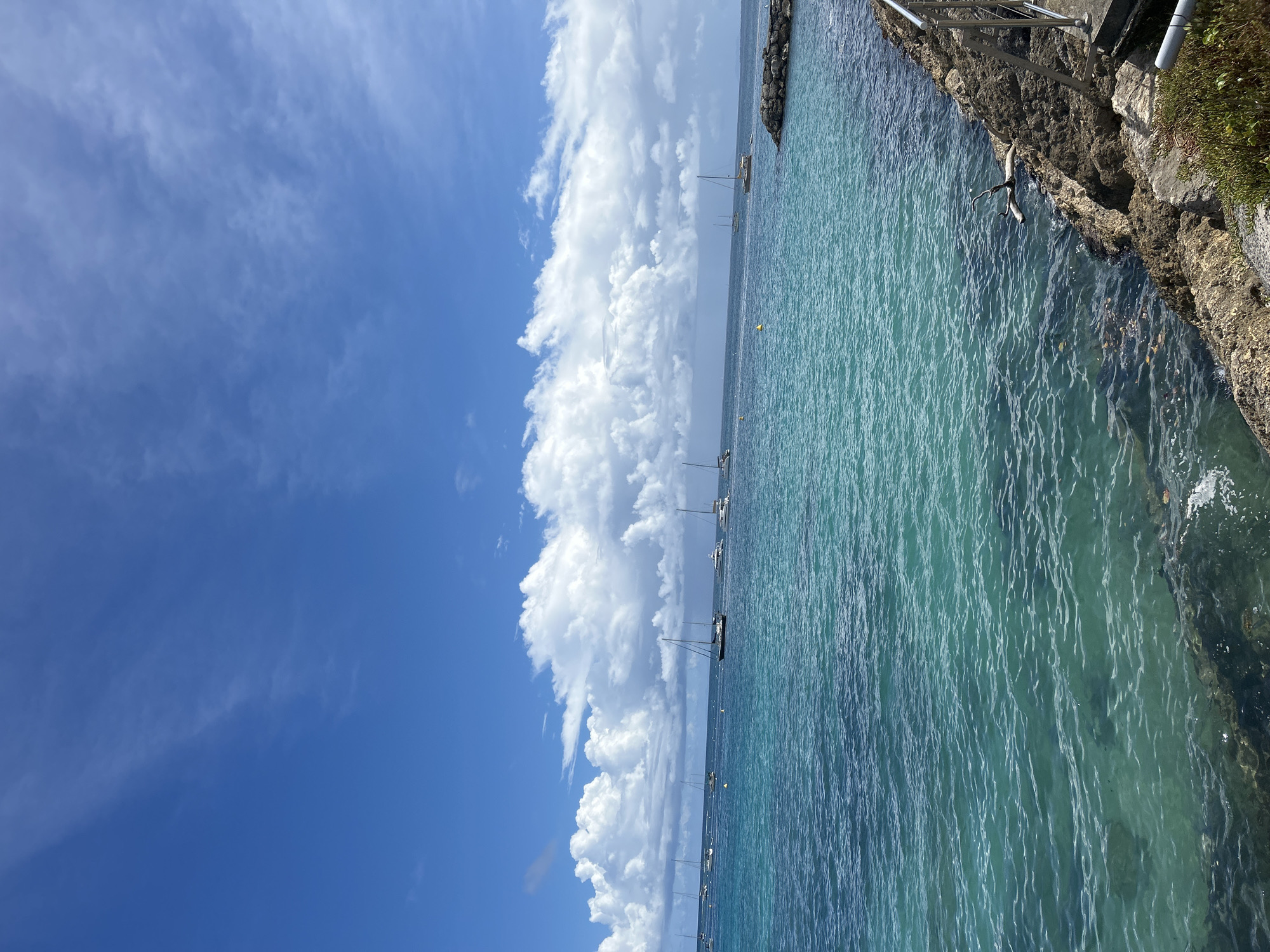 The image captures a scene at the city of Le Gosier in Guadeloupe, France. The view encompasses the bright blue sky with scattered, puffy white clouds that dominate the left portion of the image. Transitioning to the right, there's a body of water that fills the rest of the frame. Sailboats are lined in the ocean with yellow buoys, indicating a marina or mooring area. The water gradually darkens and reflects with the coastline where rough textured rocks form a barrier. There is a dark staircase with a rail in the upper right-hand corner on top of rocks. The overall view suggests a serene coastal location on a bright, sunny day.