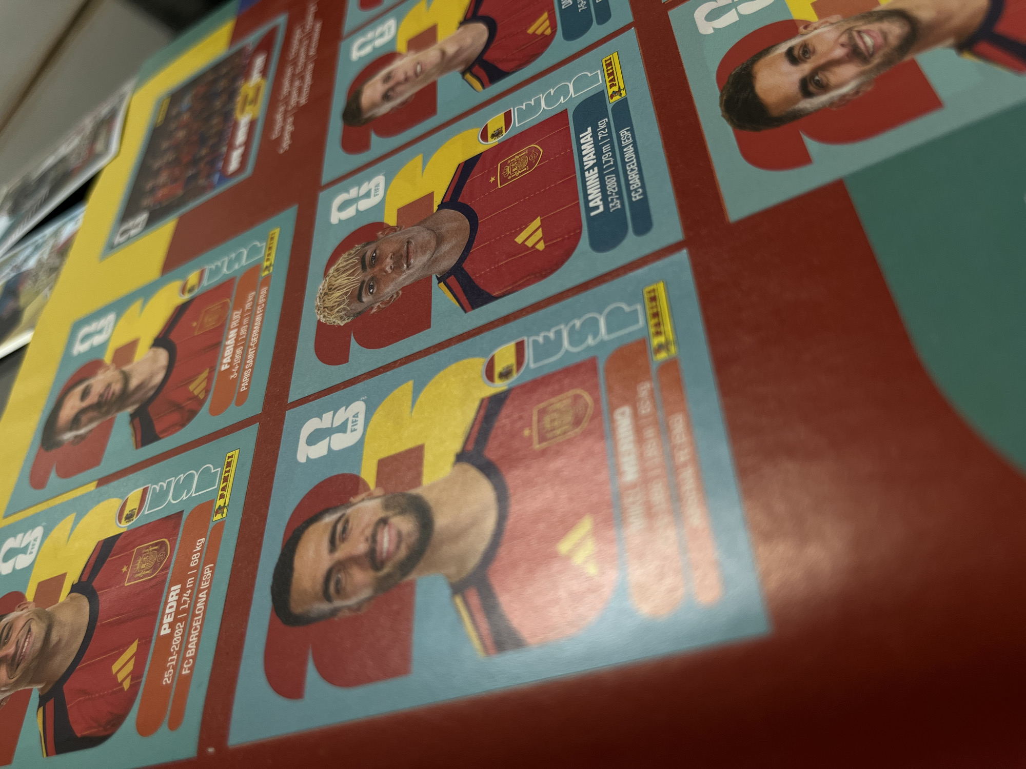 The image shows a close-up view of a Panini sticker album page. The stickers feature images of soccer players. Specifically visible are stickers of Pedri, Fabian Ruiz, Mikel Merino, and Lamine Yamal, all players for the Spanish national team and FC Barcelona. Each sticker includes the player's name, birthdate, height, weight, and club affiliation. The environment appears to be indoors on a flat surface where the sticker album is placed.