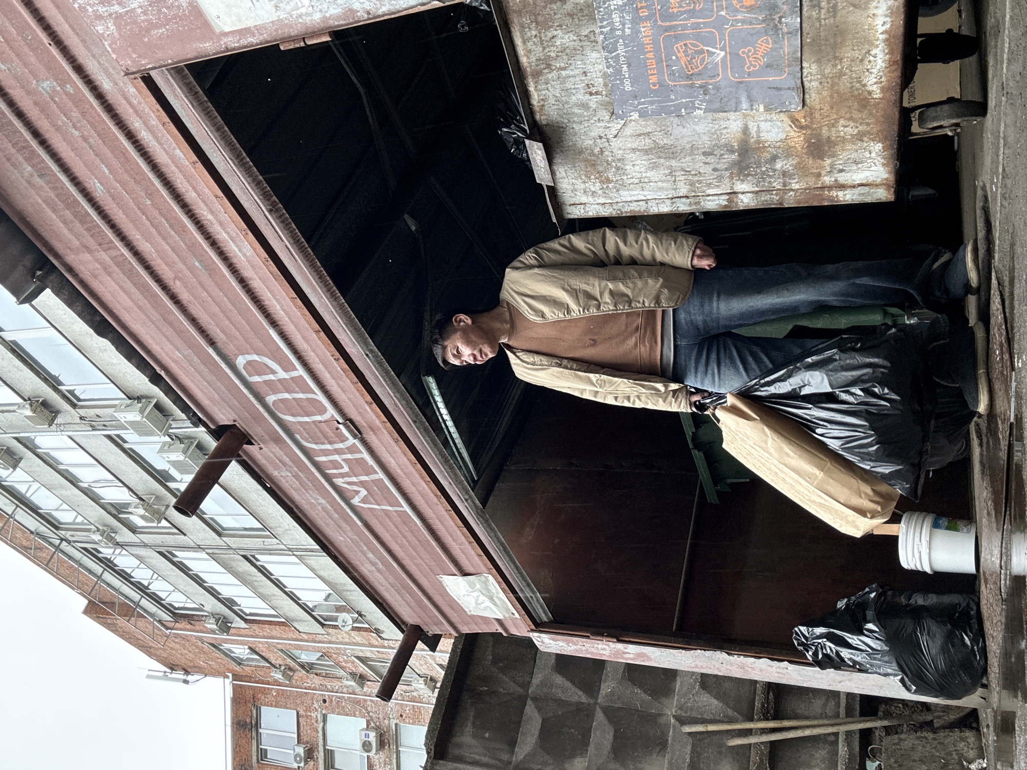 A man stands in a dumpster area in Khimki, Russia. He is wearing a beige jacket and jeans, and is holding a brown paper bag. The dumpster area is enclosed by a rusty metal structure with the Russian word "МУСОР" (meaning "trash") painted on it. Behind the dumpster area, there is a brick building with multiple windows and air conditioning units. The surrounding environment appears urban and slightly rundown. There are black trash bags and a white bucket near the man.