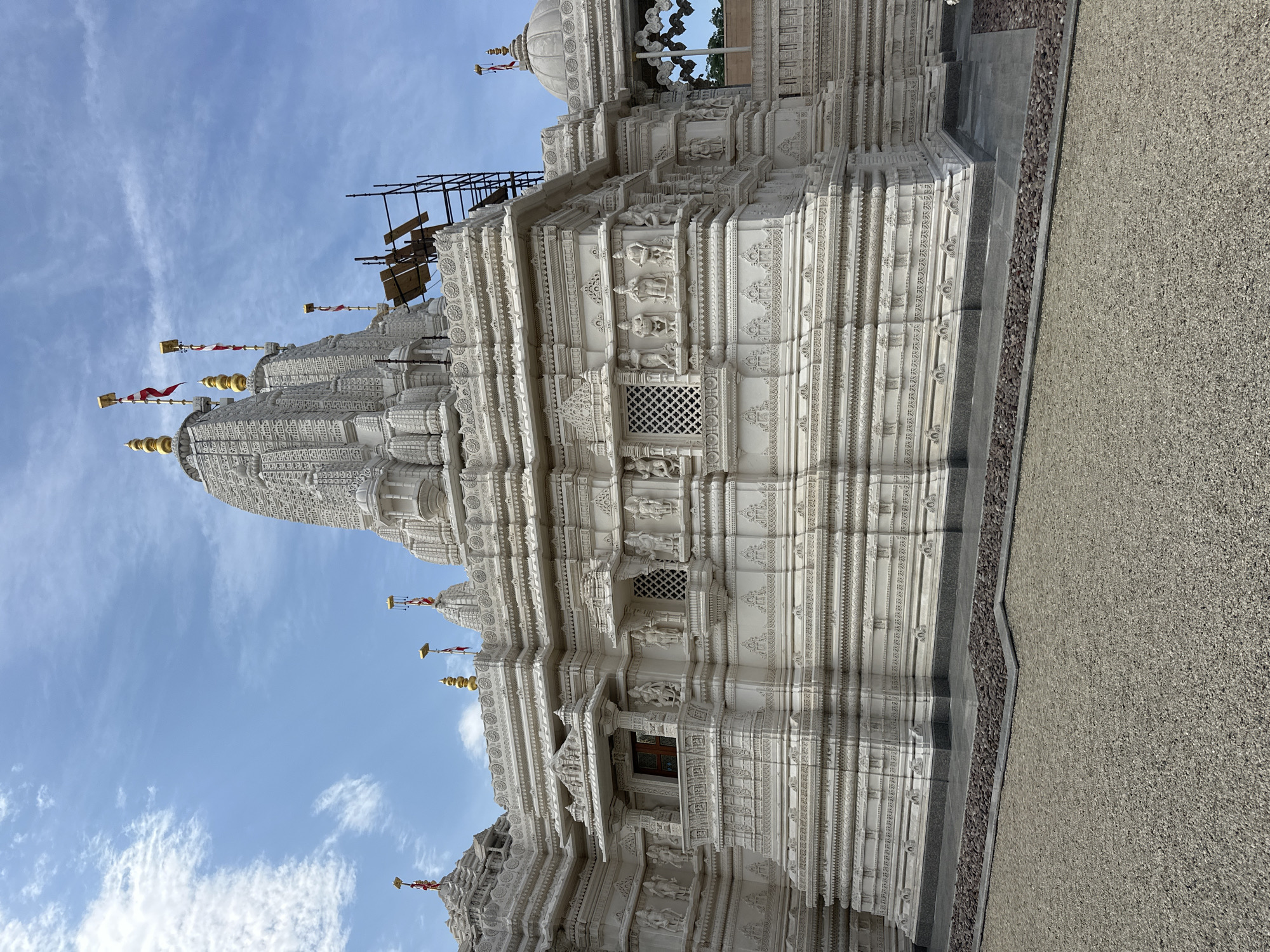 The image shows a white, intricately carved Hindu temple in Bartlett, Illinois, United States of America. The temple has multiple tiered roofs topped with golden spires and flags. The facade is adorned with numerous sculptures of deities and decorative patterns. Scaffolding is visible on the right side of the building, suggesting ongoing construction or maintenance. The temple is surrounded by a gravel courtyard. The sky is blue with scattered white clouds.