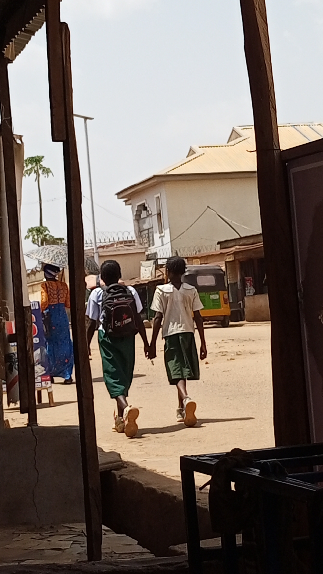 Two young individuals in school uniforms are walking hand-in-hand on a dirt road in Sabon Tasha, Nigeria. The individual on the left has a backpack. A woman carrying something on her head walks in the opposite direction. A three-wheeled vehicle is visible in the background. The scene is framed by wooden structures.
