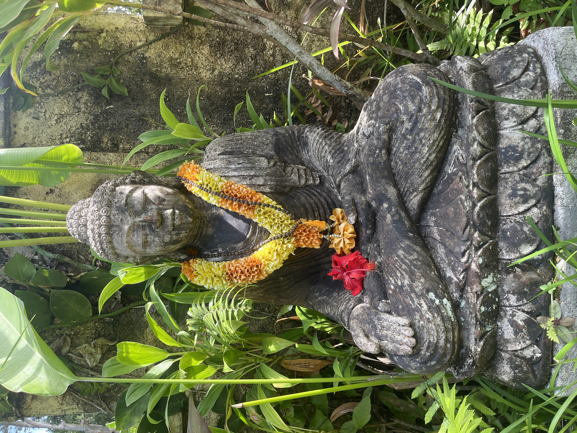 An outdoor, eye-level, medium shot captures a weathered stone statue of Buddha seated in a cross-legged meditation pose. The statue is adorned with a vibrant, layered lei of yellow and orange flowers, and a single red hibiscus rests on its lap. Lush green foliage and a textured, moss-covered wall form the backdrop. The scene is situated amidst a garden setting, likely in Ubud District, Indonesia, implied by the tropical flora and Balinese-style stone work. The lighting suggests it is daytime, with shadows indicating the sun's position.