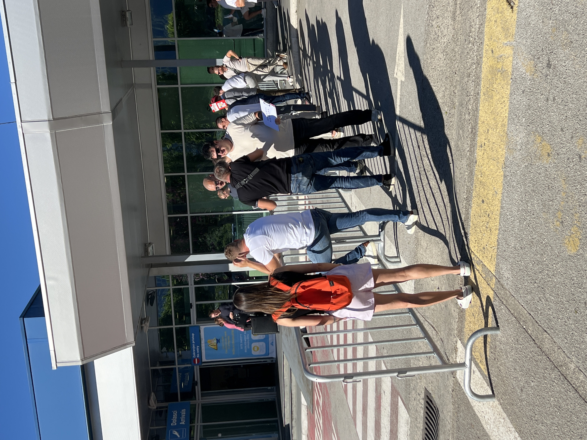 The image depicts an outdoor scene in front of an airport arrivals building, identified as being located in Mrčevac, Montenegro. A queue of people, predominantly male, are lined up outside the building under a white canopy. Some individuals are holding papers and what appears to be a red box. A young woman with an orange backpack stands facing away from the camera, near a silver barrier. The ground is paved and marked with yellow and white lines. The building's facade features large windows reflecting trees. The sky is clear and blue.