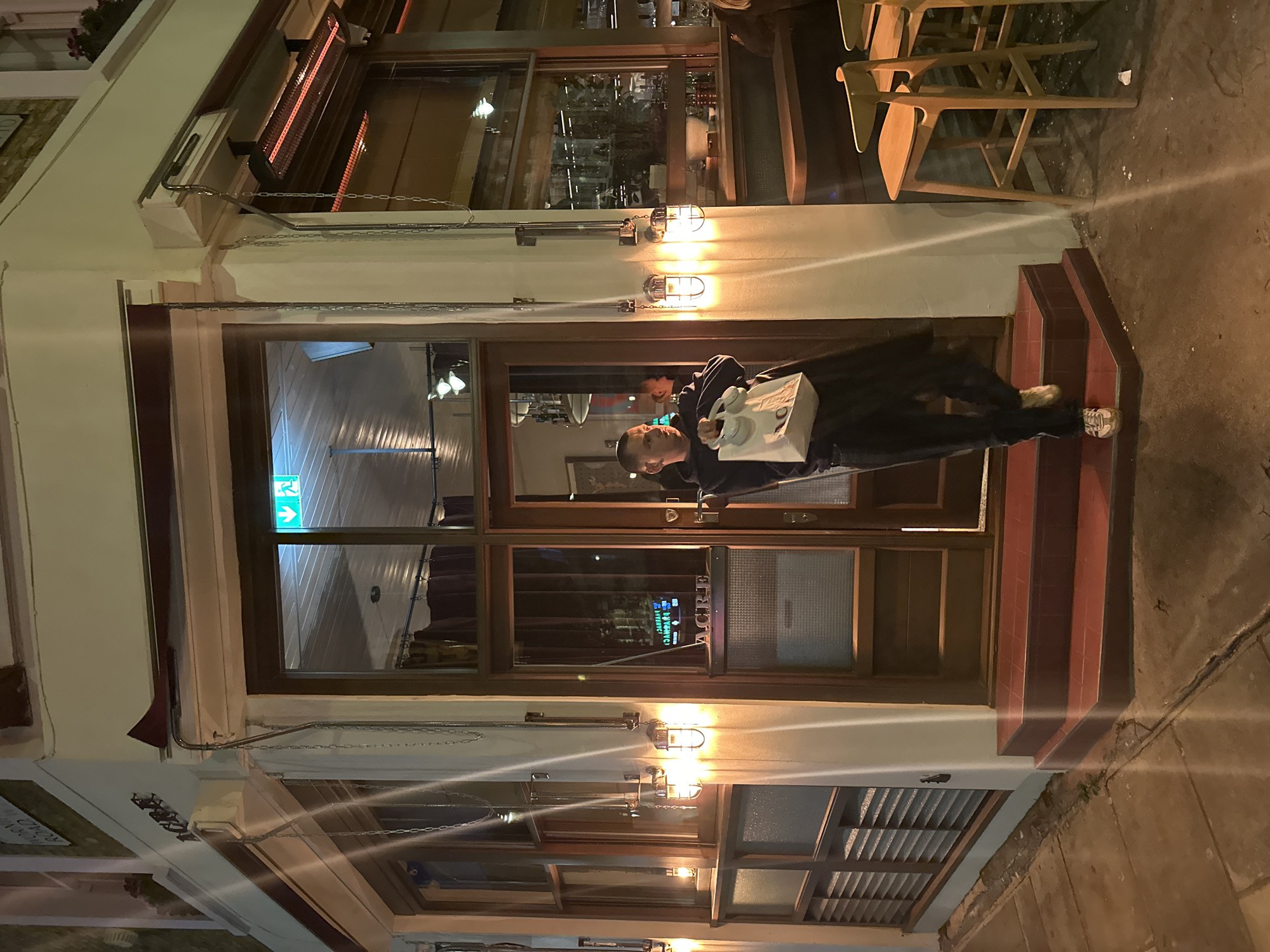 A man wearing a dark jacket and light-colored sneakers stands on a set of tiled steps in front of a building at night. He holds a white box with headphones. The building has large windows and a wooden door with a sign that reads "ACRE." Above the windows are lights and an awning. A row of wooden chairs is visible on the right side of the image. The location is likely London, United Kingdom, given the style of the building and street.