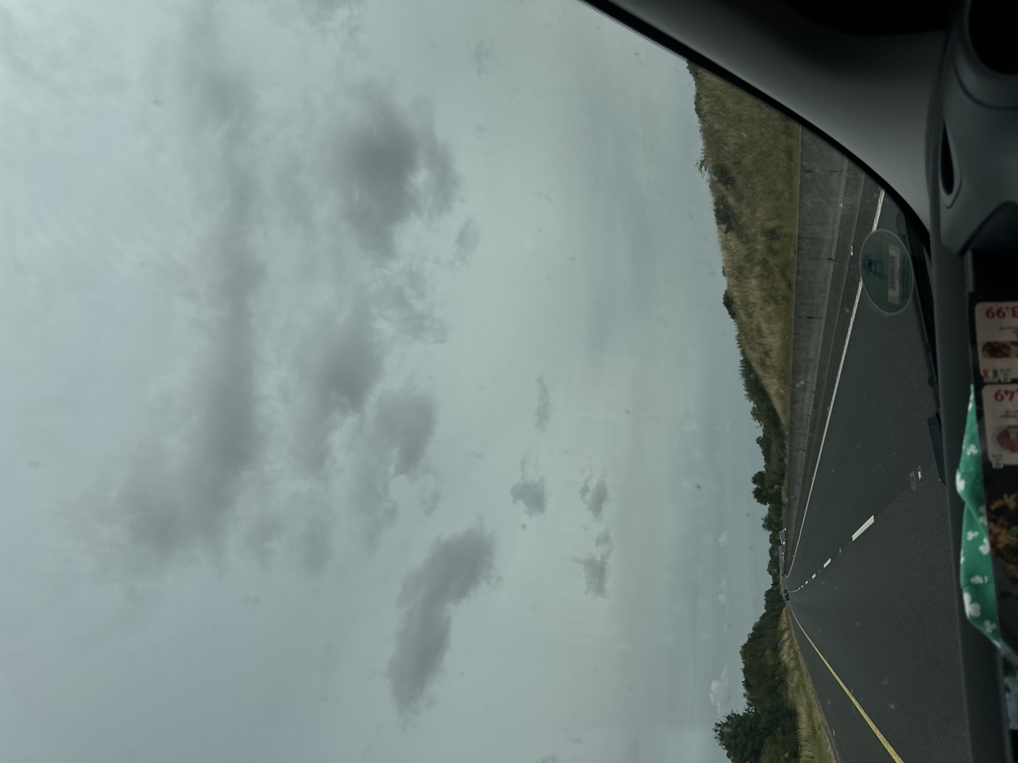 The image shows a view from inside a vehicle driving on a highway. The sky is overcast with scattered grey clouds. The highway has multiple lanes separated by white and yellow lines. The roadside is lined with green trees and grassy embankments. In the foreground, part of the vehicle's dashboard and windshield are visible, with some items on the dashboard. There are no people or specific landmarks identifiable to pinpoint a precise location in Leighlinbridge, Ireland, beyond it being a highway setting within or near the town.
