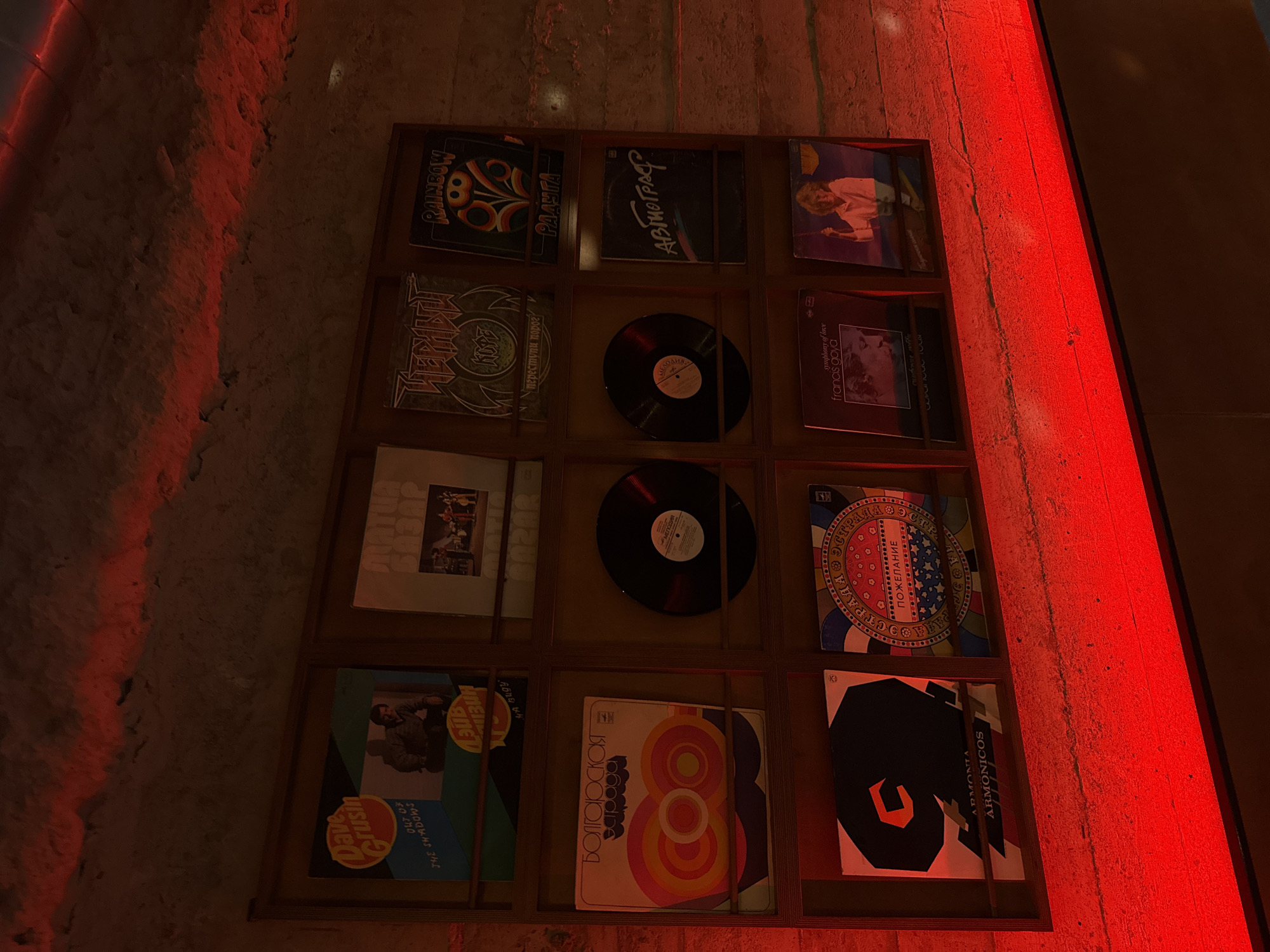 The image displays a wall of vinyl records, likely in a dimly lit establishment, possibly a bar or cafe, with prominent red lighting casting a glow on the scene. The records are arranged in a grid of three rows and three columns, with some shelves containing multiple records. The records themselves are varied, featuring different cover art and titles.
In the top left, a record titled "Dave Grusin" with "Out of the Shadows" is visible. To its right, another record appears to have a band performing on stage. The middle row on the left features a record with a psychedelic, circular graphic and Cyrillic text that reads "БОЛГАРСКАЯ ЗАРАГА". Next to it, a black vinyl record is displayed. In the top middle, a record with a stylized, gothic font reads "ЧЕРНЫЙ КЛЮЧ". To its right, a record with the title "RAINBOW" and a floral motif is visible. The middle right section shows a record with the title "АВТОГРАФ" in a flowing script. The bottom row on the left has a record with a large black numeral "9" and "ARMONIA ARMONICOS". To its right, a colorful record with Cyrillic text that says "ПОЖЕЛАНИЕ" is displayed. The bottom right section features a record with a woman on a swing.
The red lighting suggests a deliberate atmosphere, perhaps intended to create a moody or vibrant ambiance. The rough texture of the wall behind the records hints at a rustic or industrial interior design. The overall impression is that of a curated collection of music, presented as art.
