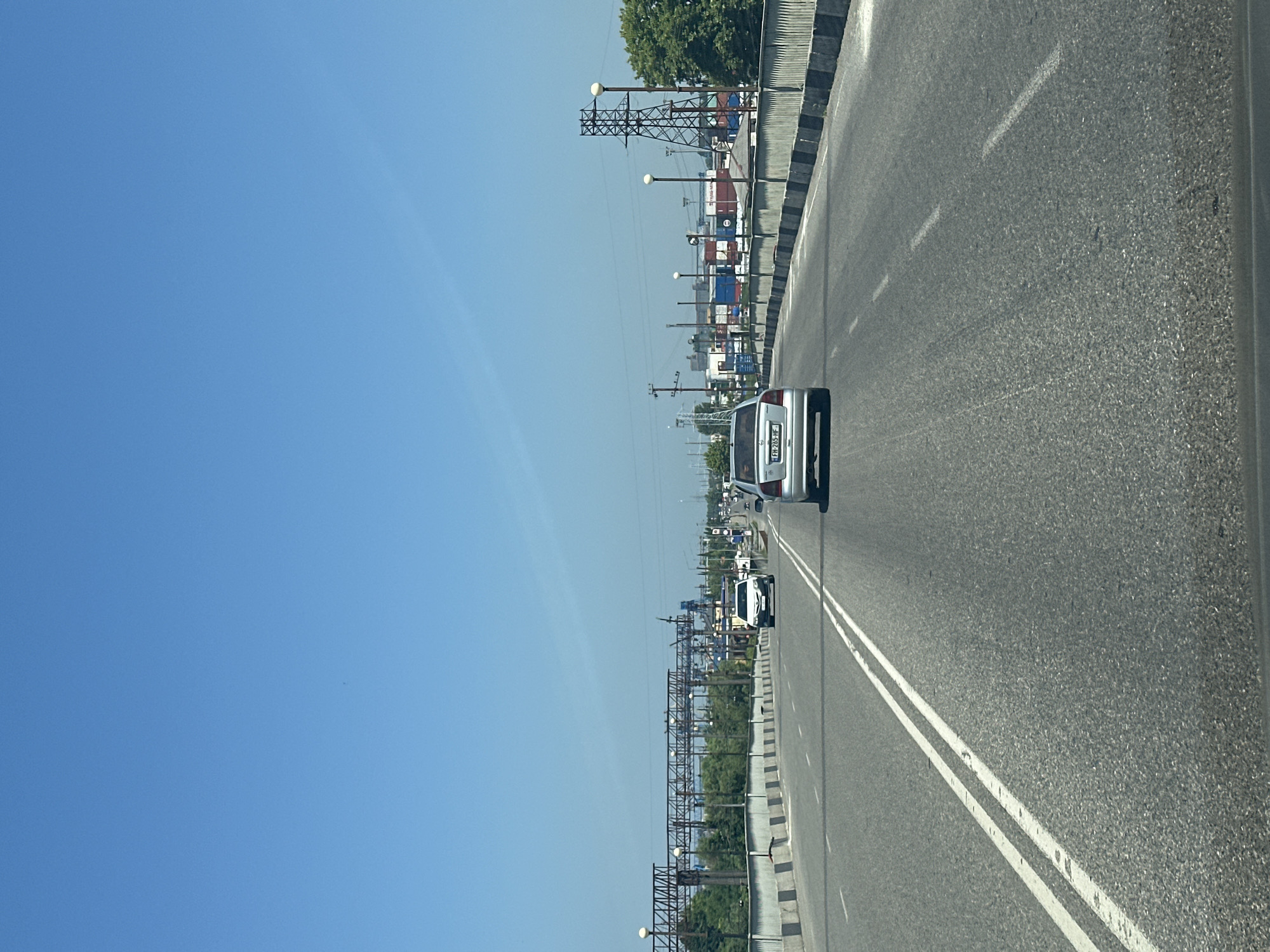 A bright, clear blue sky is visible above a multi-lane asphalt road. The road appears to be on an elevated structure, possibly a bridge or overpass, with a concrete barrier lining the right side and a metal railing along the left side. Several cars are on the road, traveling in the same direction. The car in the foreground, on the right, is silver with a visible license plate. To the left, a white car is further ahead. In the distance, industrial structures, including large metal frames and shipping containers, are visible, suggesting proximity to a port or industrial area. Green trees line the left side of the road in the background. Power lines and utility poles are also present. The overall scene depicts a sunny day in an urban or industrial setting, likely the city of Poti, Georgia, given its known port and industrial activities.
