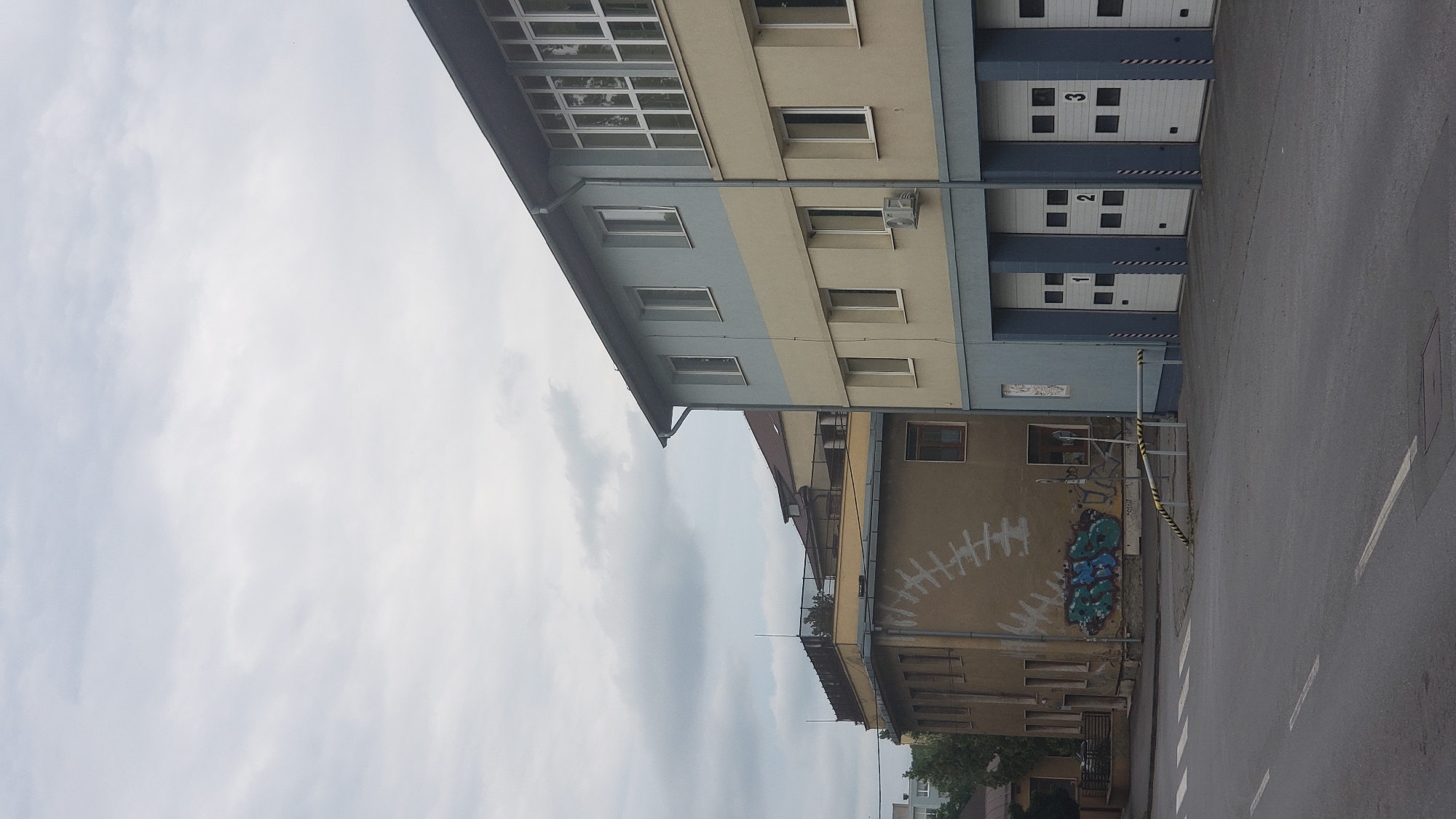 The image depicts an exterior view of a building with several garage bays, numbered 1, 2, and 3, in the city of Prešov, Slovakia. The building has a light blue and beige facade. To the left of this building is another, older structure with a light brown facade, which features graffiti on its lower portion. The scene is captured on a cloudy day, with the sky overcast. The foreground shows a paved road with white lane markings. A striped barrier pole is visible in front of the older building. The image appears to be taken from a street level perspective, looking towards the buildings. There are no people visible in the image.