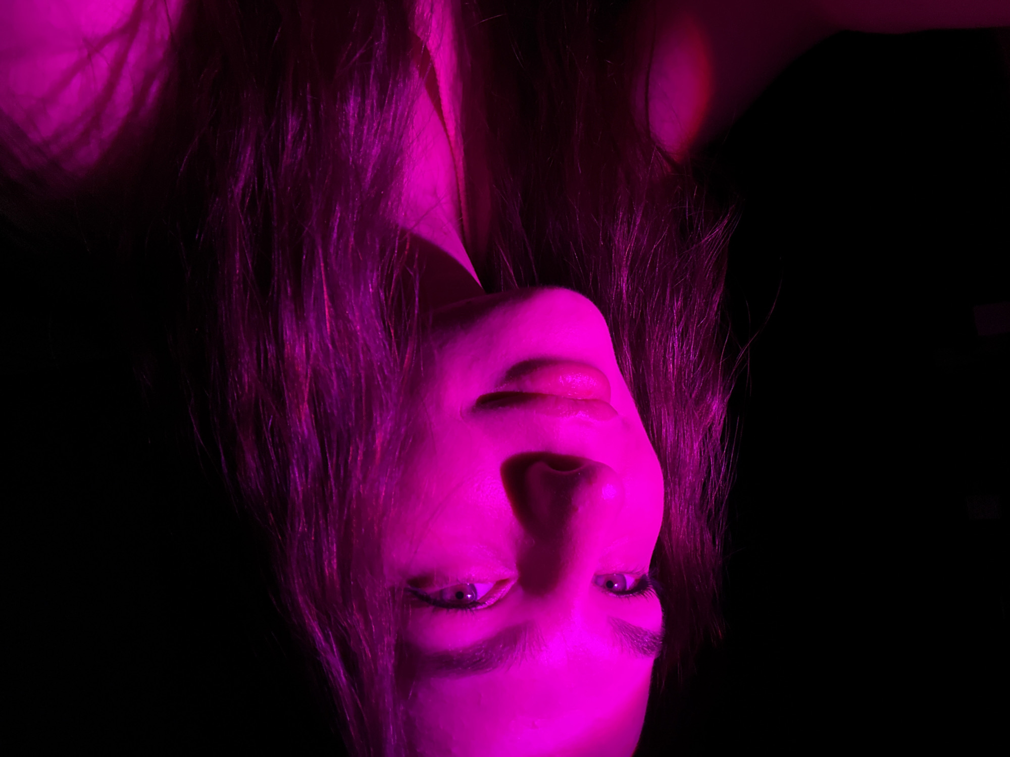 A female figure with long hair is illuminated with a strong, monochromatic pink light against a predominantly black background. The figure is oriented upside-down relative to the frame. The face is upturned toward the light source, highlighting features such as eyes, nose, and lips. The surrounding environment is largely obscured by darkness, making it difficult to determine the location in Chaikovsky, Russia.