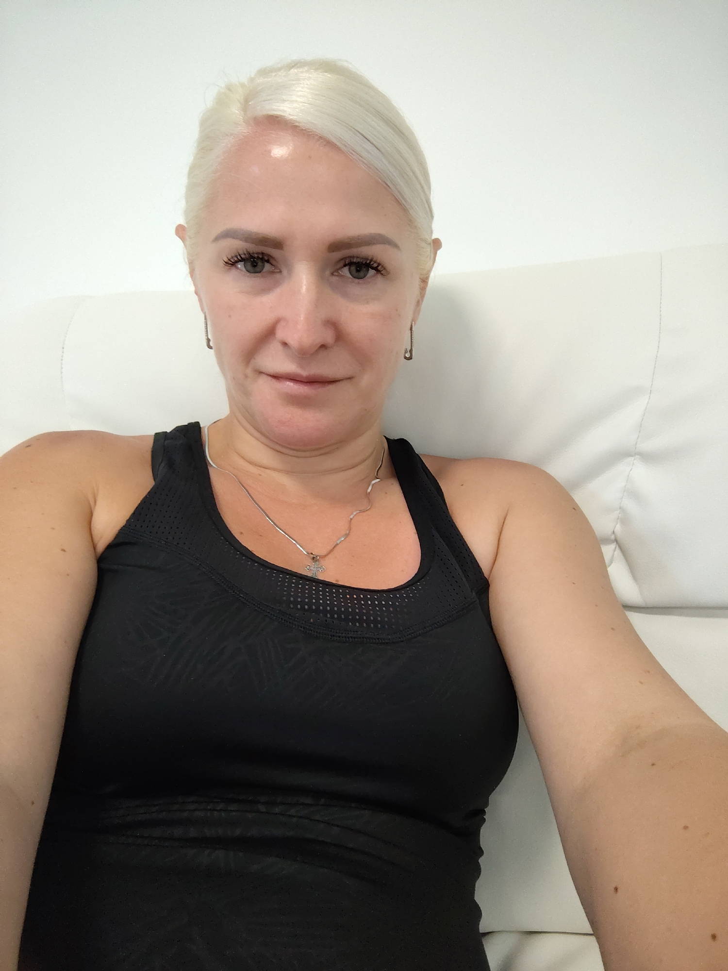 A blonde woman with her hair pulled back is taking a selfie indoors. She is wearing a black athletic tank top and a silver necklace with a cross pendant. She is sitting on a white padded surface, likely a couch or chair. The background is a white wall and the white padded surface. There are no discernible details indicating the location is specifically in Chaiky, Ukraine.