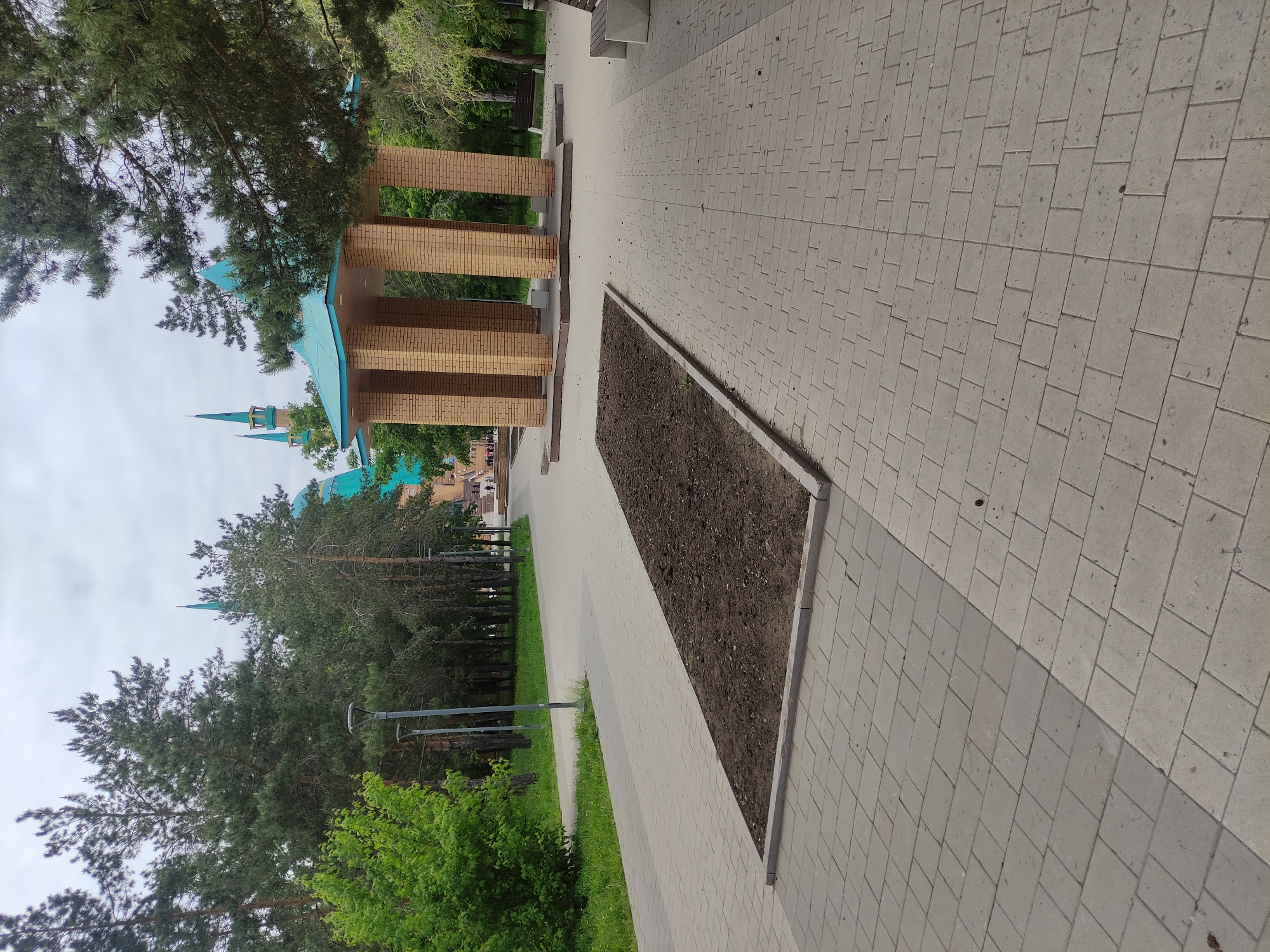 The image shows a park in Pavlodar, Kazakhstan. A tiled walkway leads the viewer's eye towards a light-brown brick structure with a turquoise roof, located in a park. The park has tall green trees. The walkway has a flower bed located beside it on the lower side of the image. Another walkway runs next to the brown brick structure. The sky is cloudy. The structure appears to be a gazebo. The image is taken at eye level. There is no one visible in the image.