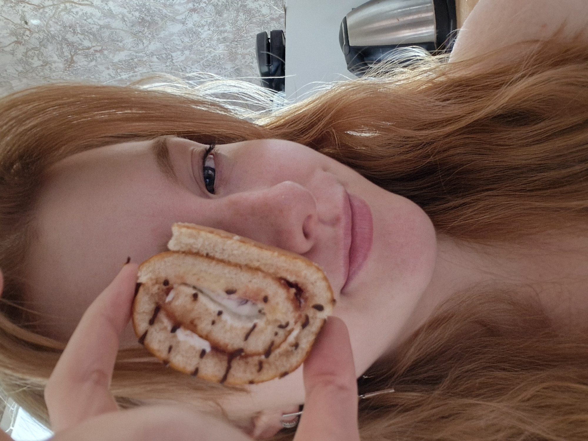 A woman with long, wavy red hair is holding a piece of Swiss roll close to her face. The Swiss roll has a light-colored cake base with a white cream filling and a drizzle of dark chocolate on top. The woman has fair skin, freckles, and blue eyes. In the background, there is a kitchen with a white countertop and some appliances. The location appears to be indoors. There is no visual information to ascertain its location in Yekaterinburg, Russia.