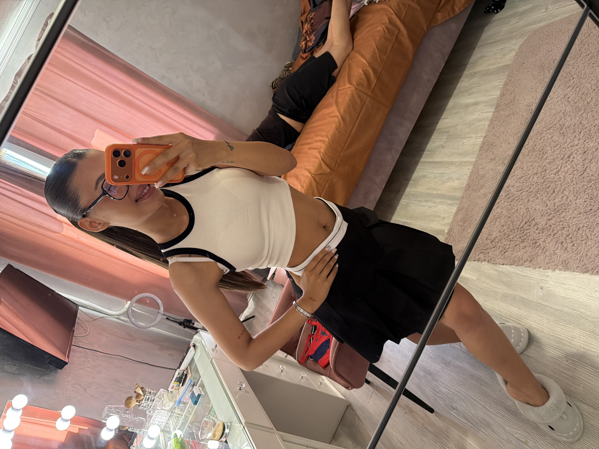 A young woman, appearing to be in her late teens or early twenties, is taking a mirror selfie. She is wearing a white tank top with black trim, a black pleated skirt, and grey slippers. She has her hair pulled back in a high ponytail, and she is wearing black-rimmed glasses. She is holding an orange smartphone in her right hand, angled to capture the mirror's reflection. Her left hand is resting on her hip. In the background, a makeup vanity with lights is visible, as is a pink curtain and a bed with a person lying on it. The scene is likely an interior residential setting. There is no information in the image to suggest a specific location in Burgas, Bulgaria.
