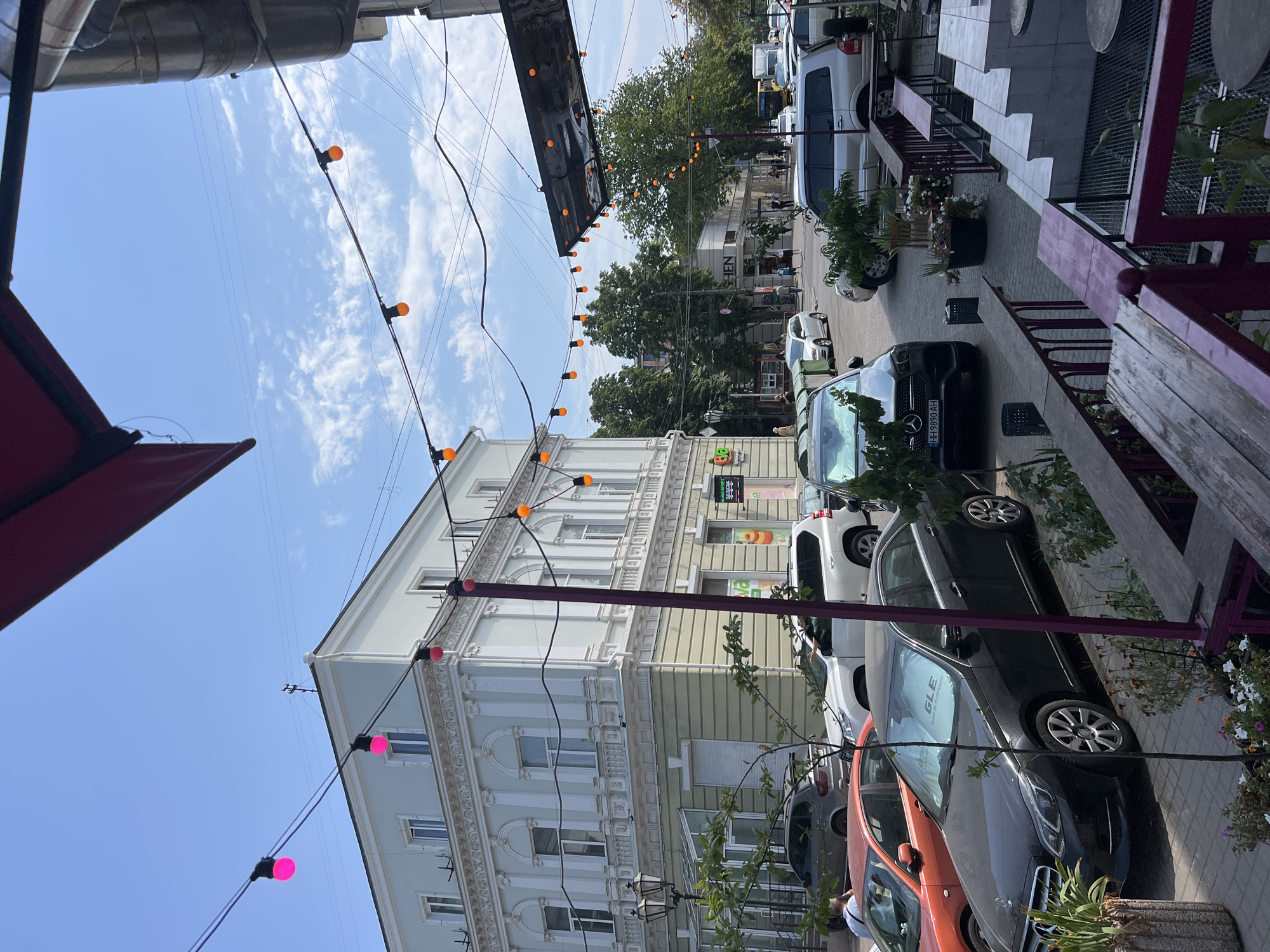 This is a photograph taken on a sunny day in Odesa, Ukraine, likely on a street with a mix of residential and commercial buildings. In the foreground, there is outdoor seating for a cafe, with wooden tables and benches, and a railing made of dark wood and metal. String lights with pink and orange bulbs are strung across the street, adding a festive atmosphere.
The street is lined with parked cars, including a black Mercedes-Benz sedan in the foreground, an orange sedan, a white SUV, and a silver SUV further down. Pedestrians can be seen walking on the sidewalk and crossing the street.
The architecture of the buildings is notable, with a light blue building featuring ornate detailing around the windows and a decorative balustrade. A sign on one of the buildings reads "JEN". Green trees and plants are scattered throughout the scene, providing pops of color against the urban backdrop. The sky is a clear blue with scattered white clouds. The overall impression is a vibrant and lively street scene in a European city.