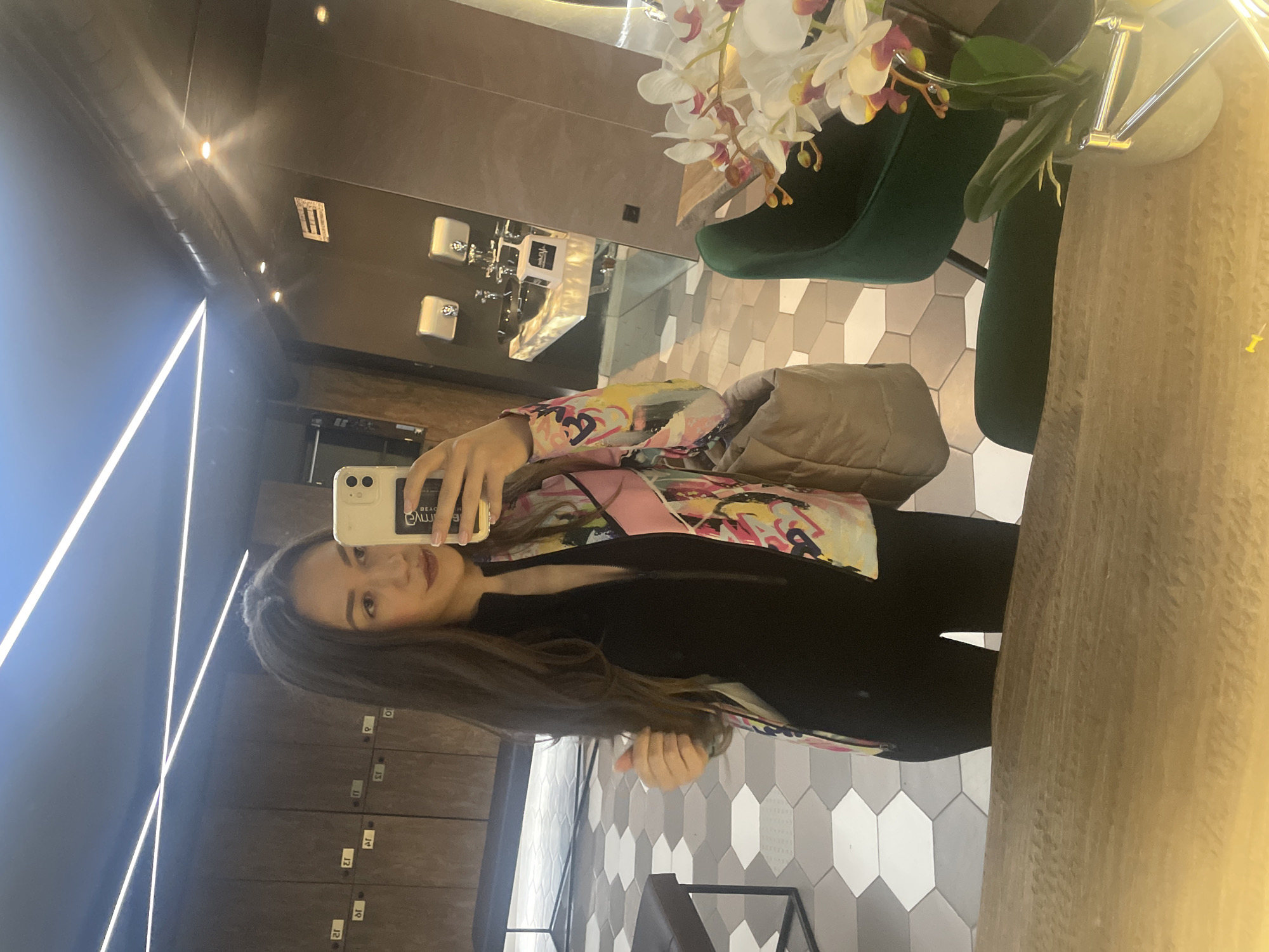 A young woman is captured in a mirror selfie within a modern indoor setting, likely a changing room or stylish lounge area in Sofia, Bulgaria. She has long, dark brown hair and a subtle smile, looking directly into the camera. She is dressed in a black top or dress, overlaid with a vibrant, multi-colored jacket featuring abstract designs in shades of pink, yellow, blue, and black. A light-colored smartphone, held up for the selfie, displays "BEAC emwA" in reverse on its screen. A beige, possibly quilted, shoulder bag is visible on her left side.
The background reveals a contemporary space with dark walls and a floor adorned with geometric hexagonal tiles in white and various shades of gray. Modern linear LED lights create striking reflections on the ceiling and walls. To the right, a display of white and pink artificial orchids sits in a vase on a light-colored countertop, with a dark green upholstered chair partially visible behind them. To the left, a row of dark lockers with reversed silver numbering (1 through 15) is reflected in the mirror. A small white label with a reversed "1" and another dark label, possibly "F" or "IT" reversed, are visible on the reflective surfaces. The overall atmosphere is clean and well-lit by artificial light.