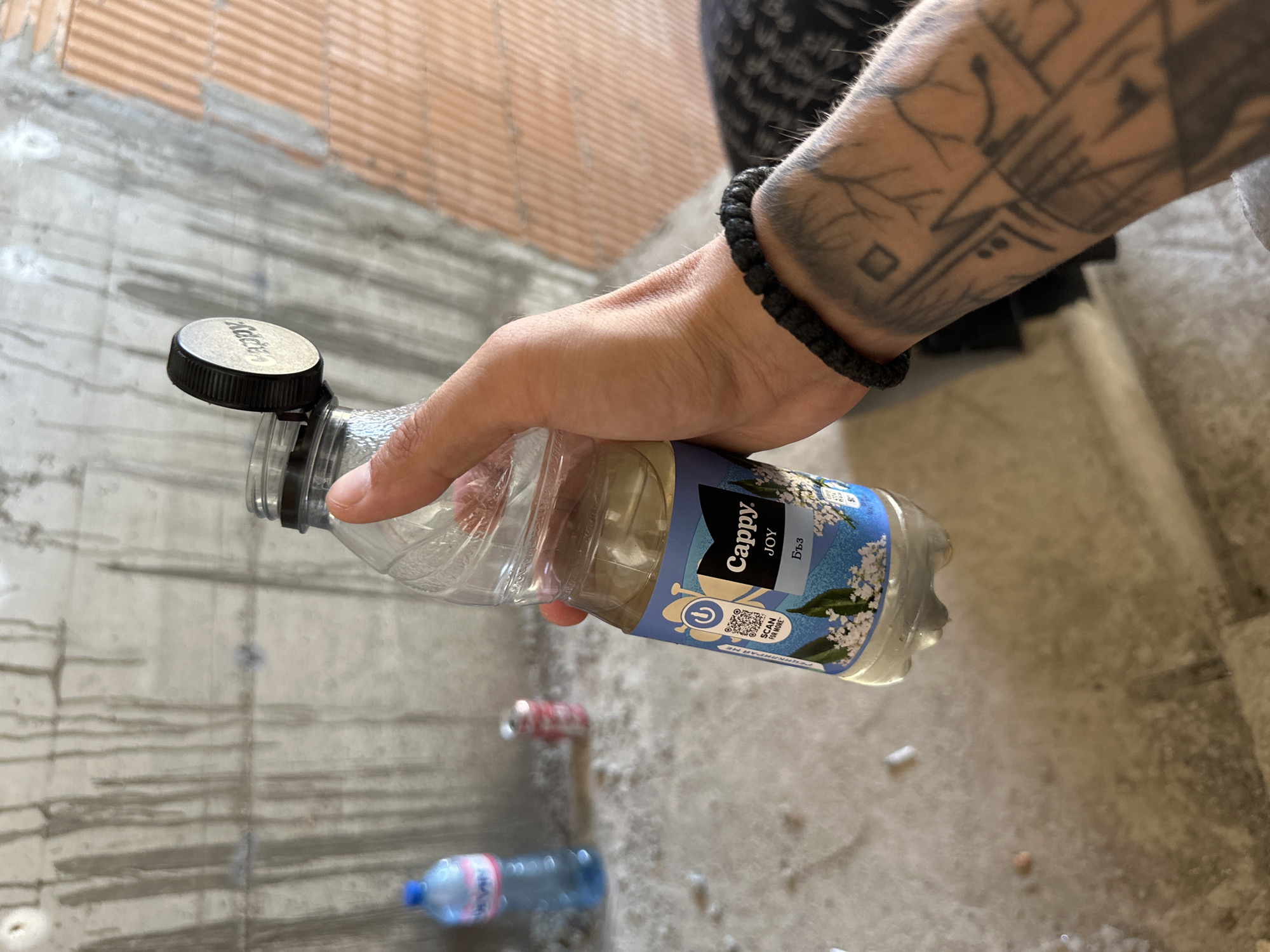 The image captures a close-up view of a hand holding a clear plastic bottle of Cappy Joy. The beverage inside is a pale yellow color. The bottle has a blue label with white text and imagery of white flowers. The cap of the bottle is black and slightly ajar. The hand holding the bottle has a black beaded bracelet on the wrist and a tattoo on the forearm featuring geometric shapes and tree-like branches. In the background, out of focus, are a blue bottle of water, a red and white can, a concrete wall with vertical stains, and orange brickwork. The ground appears to be concrete with debris.
The scene is consistent with an unfinished or construction site, potentially in the city of Stara Zagora, Bulgaria, given the presence of Cyrillic script on the beverage label, specifically "ББЗ" which is a common abbreviation for the municipality of Stara Zagora. The overall impression is that of a brief pause during work or an exploration of a site.