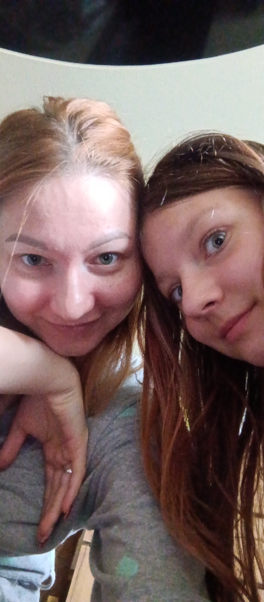 Two individuals are close together, appearing to be in a selfie. The person on the left has light brown hair and is wearing a gray sweater. A ring is visible on their left ring finger. The individual on the right has darker red-brown hair, with what appear to be thin gold tinsel strands woven into their hair. Their precise location within the city of Unknown, Unknown is not discernible from the image.