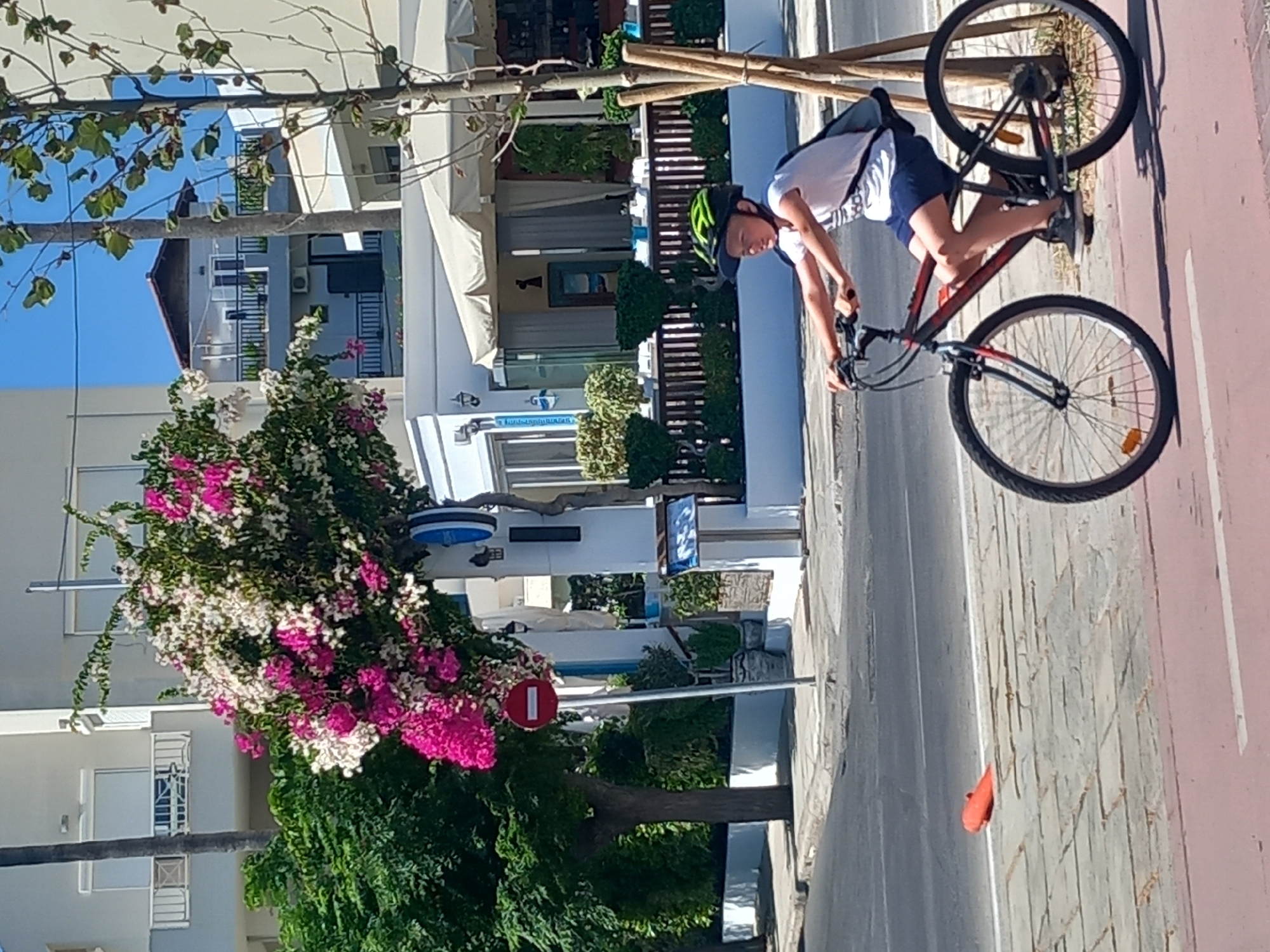 The image shows a young boy wearing a green helmet and a white t-shirt, riding a bicycle on a paved path. The path is next to a street with a red and white striped lane. The boy is riding on the right side of the image, and his bicycle is dark red and black. In the background, there are buildings, trees, and flowering bushes. The buildings appear to be in a Mediterranean style, with white walls and balconies. There is a street sign with a red circle and a diagonal line, indicating a prohibition, and a blue sign with text that is not legible. The overall scene suggests a sunny day in a town or city. The presence of bougainvillea flowers, white stucco buildings with balconies, and the general aesthetic strongly suggest the image was taken in Kos town, Greece, likely in a pedestrian or cycling area near a street.