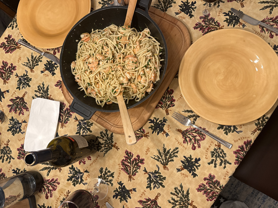 The image displays an indoor dining table set for a meal. The table is covered with a tan tablecloth featuring a repeating pattern of bundled herbs labeled "sage," "heather," and "rosemary." A large, black, round serving pan, positioned centrally on a light-wood circular trivet, contains spaghetti with shrimp and chopped green herbs. Two wooden serving utensils with heart-shaped cutouts are present within the pasta.
To the left of the pan, a dark glass wine bottle, uncorked and partially empty, stands upright. Adjacent to it, a clear glass contains red wine. A clear glass water bottle is also visible in the upper left. A folded white paper napkin is positioned near the wine bottle. Two empty, matte yellow/ochre ceramic plates are present, one in the upper right and one in the lower center-right. Each plate is accompanied by a silver fork and knife. No individuals are visible. The specific location within Los Angeles, United States of America, is not discernible from the visual information provided.