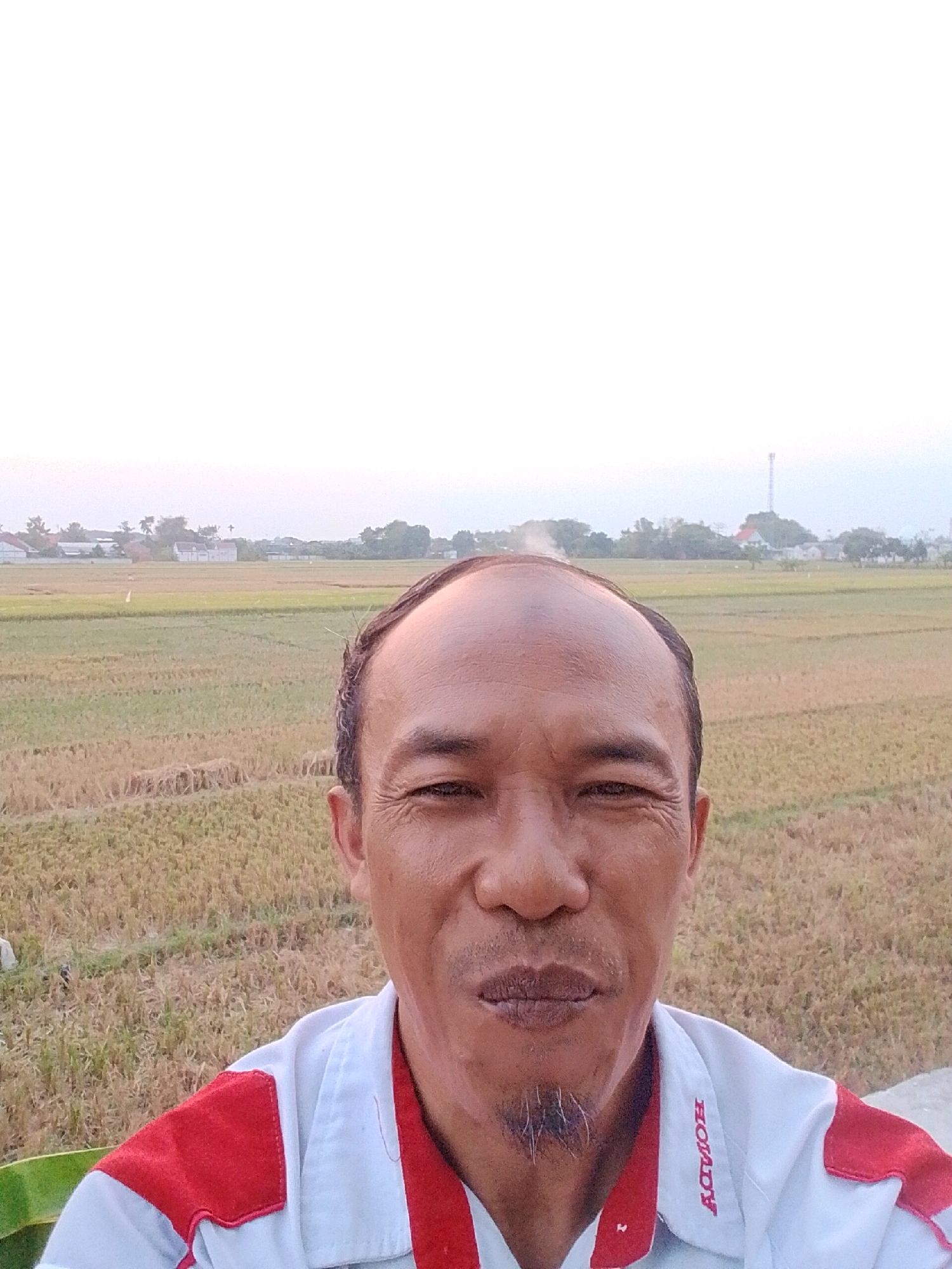 A selfie taken outdoors in Pasuruan, Indonesia shows a man in his 40s with a bald head and a goatee. He is wearing a white polo shirt with red accents. The background consists of a rice field and a few buildings in the distance. The sky is overcast.