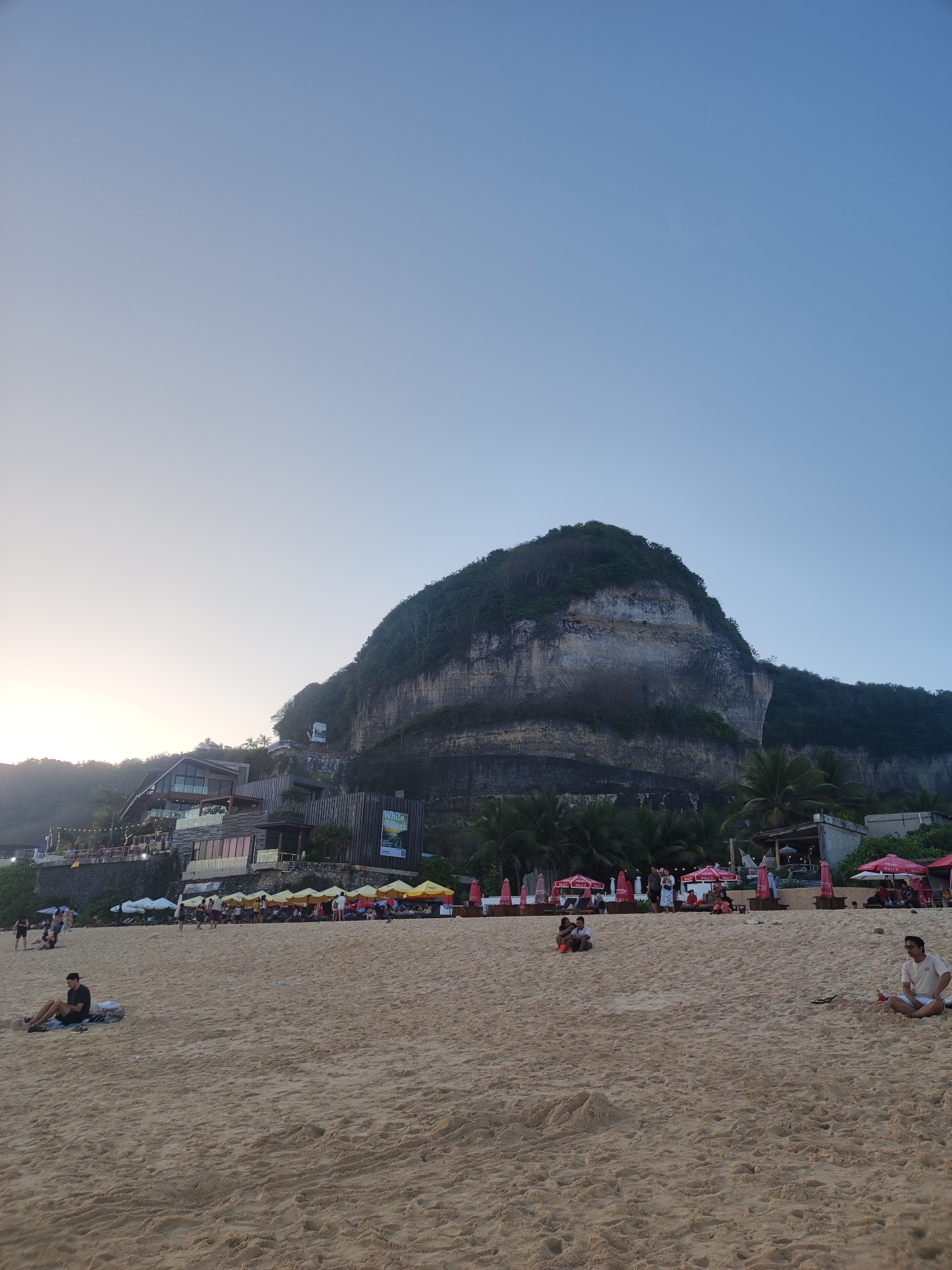 The image depicts a beach scene in Kuta Selatan, Indonesia, likely during the late afternoon or early evening. A large, vegetated cliff face dominates the background, with a multi-story building constructed into its side, featuring modern architecture with large glass windows. Numerous beach umbrellas, mostly yellow and pink, are set up on the sand, indicating a resort or restaurant area. Several people are visible on the beach, some sitting or lounging on the sand, while others are walking. The sand is a light tan color, and the sky is a clear, pale blue, transitioning to a lighter hue near the horizon where the sun is likely setting or has recently set. Palm trees are scattered along the treeline at the base of the cliff. The scene suggests a relaxed atmosphere at a popular tourist destination.