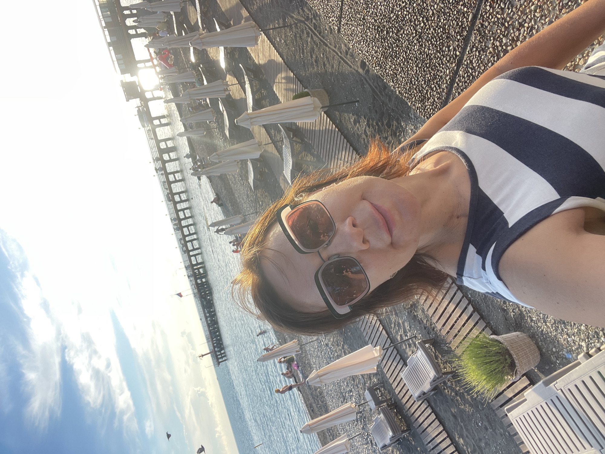 A woman with auburn hair, wearing sunglasses and a blue and white striped top, takes a selfie on a pebble beach. She is looking directly at the camera with a slight smile. In the background, the sea stretches out to the horizon under a bright, hazy sky. A wooden pier extends into the water, with people visible on it. Rows of beach chairs and closed umbrellas are set up on the sand. The sun is setting, casting long shadows and a warm glow over the scene. The location is identified as the city of Sochi, Russia.