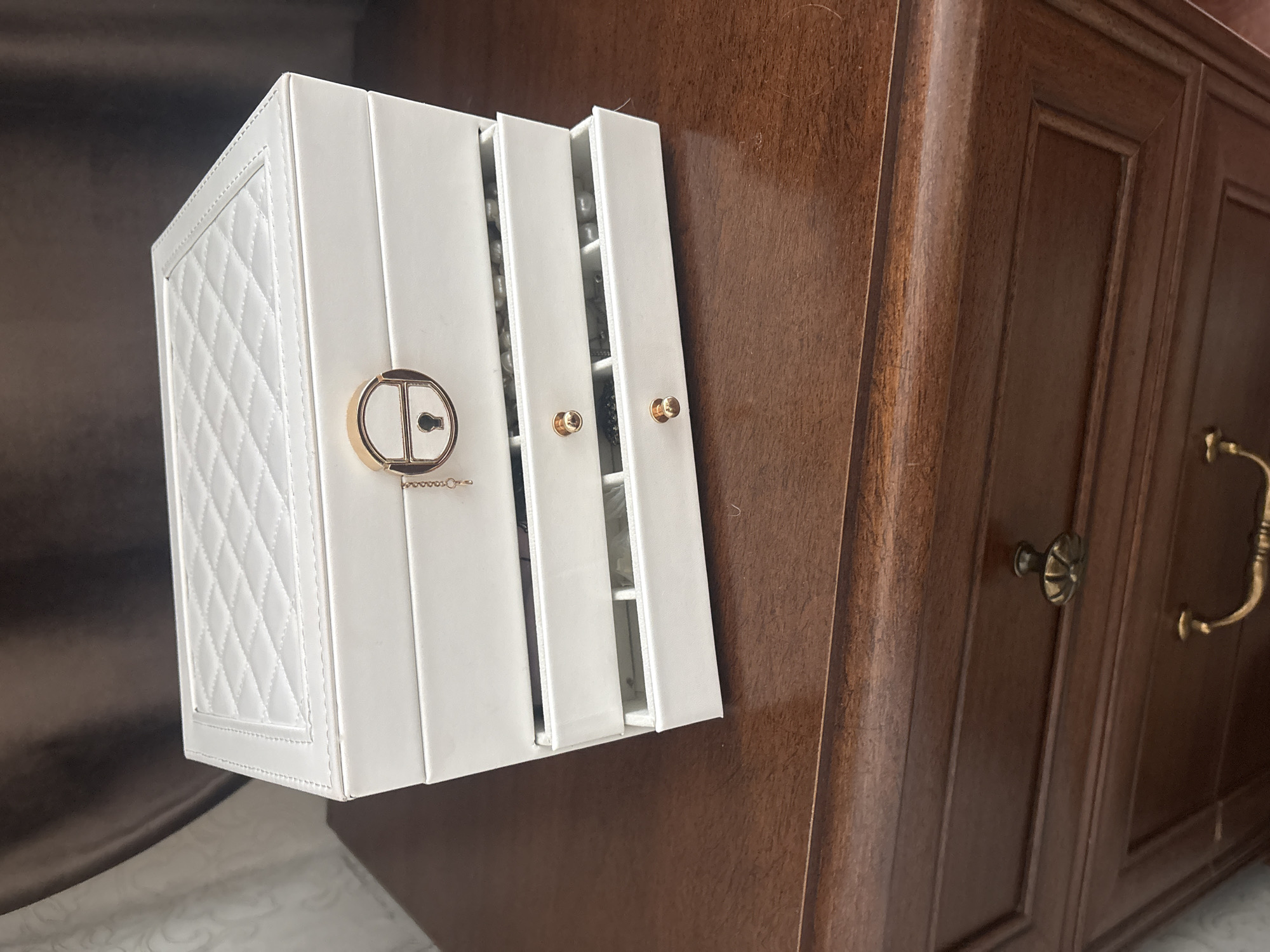 A white jewelry box with a diamond-stitched pattern sits on a wooden cabinet with ornate hardware. The jewelry box has three compartments, two of which are open, revealing what appears to be jewelry inside. The cabinet is dark brown and has a decorative design. The scene is indoors, and the lighting is somewhat dim.