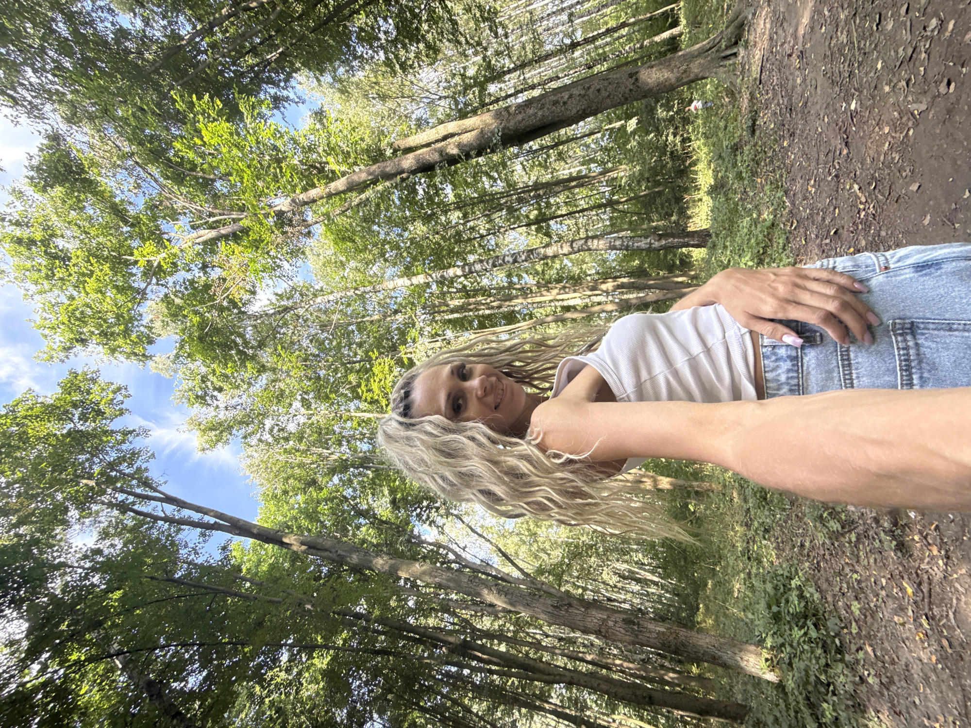 A low-angle, wide-shot photograph captures a young Caucasian woman with long, blonde, curly hair and a white tank top posing for a selfie in a forest. She is smiling and looking at the camera, with her right arm extended and her hand resting on her denim shorts. The background is filled with tall trees and greenery, characteristic of a natural forest environment. The ground is covered with dirt, leaves, and patches of grass. The sky is visible through the canopy of leaves, showing a clear blue sky with some wispy clouds. The scene appears to be set in a park or wooded area, possibly in or near Rusanovo, Russia, given the context. The lighting suggests it is daytime, with sunlight filtering through the trees.